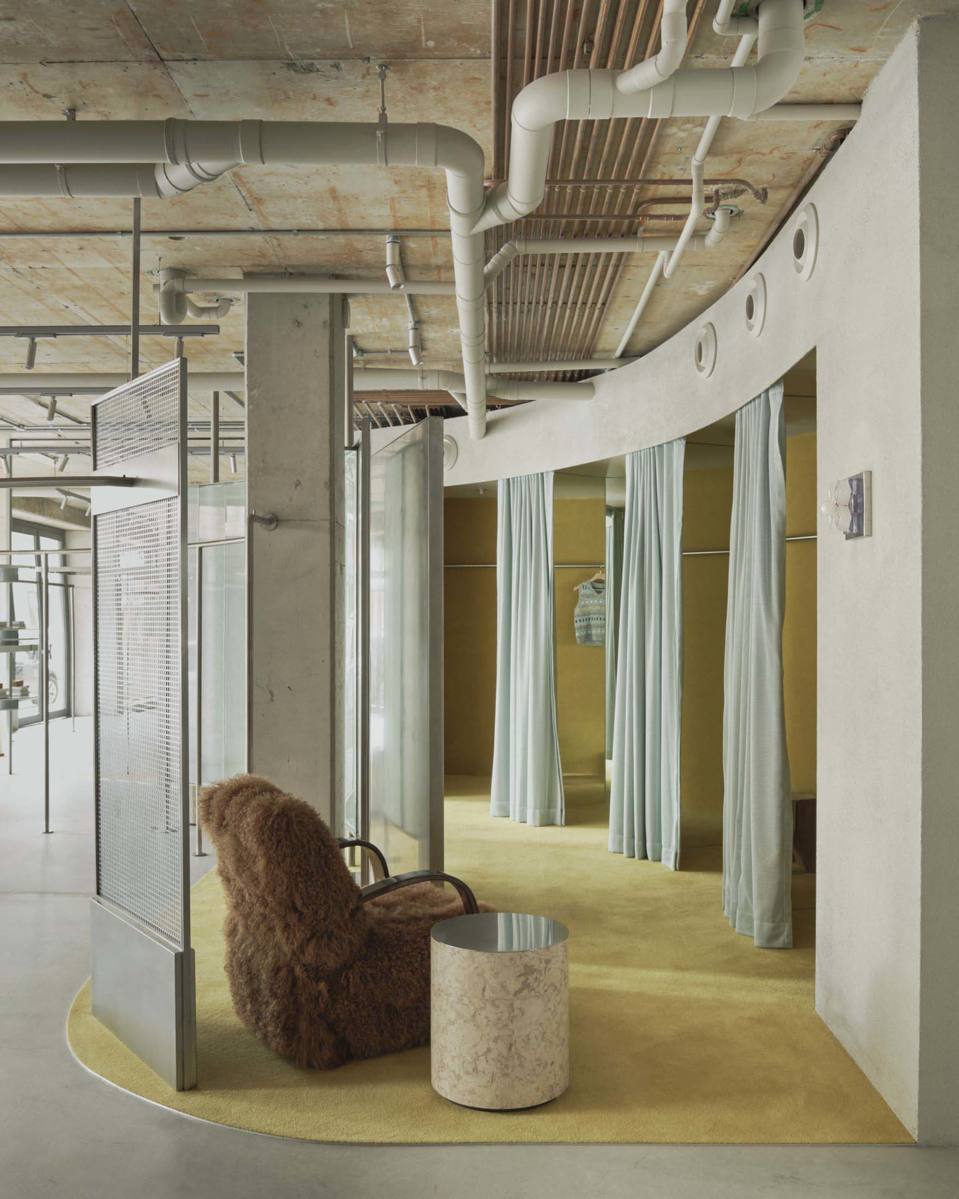 Modern fitting room area with curved concrete wall, exposed ceiling pipes, chartreuse carpet, brown sheepskin-covered armchair and marble side table