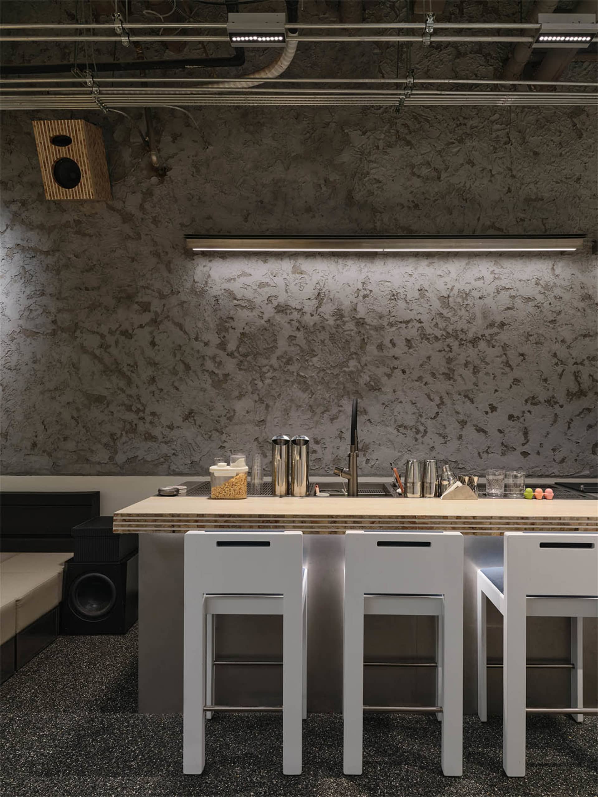 Industrial style kitchen bar counter with raw concrete wall, white bar stools, and exposed ceiling pipes.