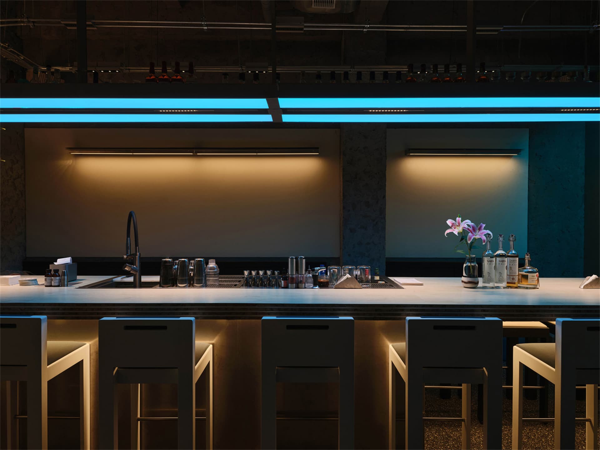 Modern industrial bar counter with four white bar stools, black faucet, and dramatic blue LED neon lighting above a concrete backdrop.