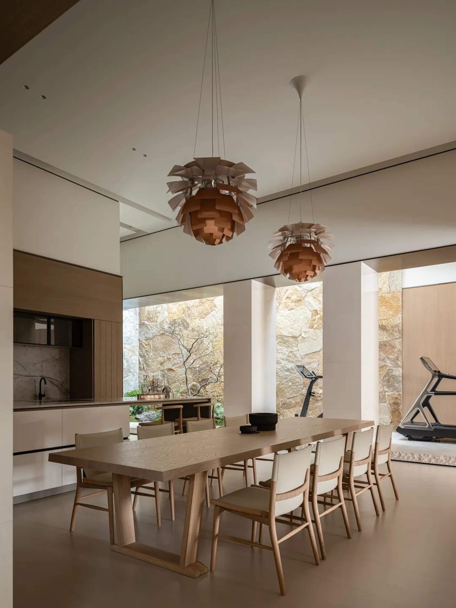 Modern minimalist dining room featuring a long solid oak table, eight matching oak dining chairs with light upholstery, two large wooden artichoke pendant lights, white walls, and a dramatic natural stone feature wall visible through large openings, with a kitchen counter and treadmill in the background.
