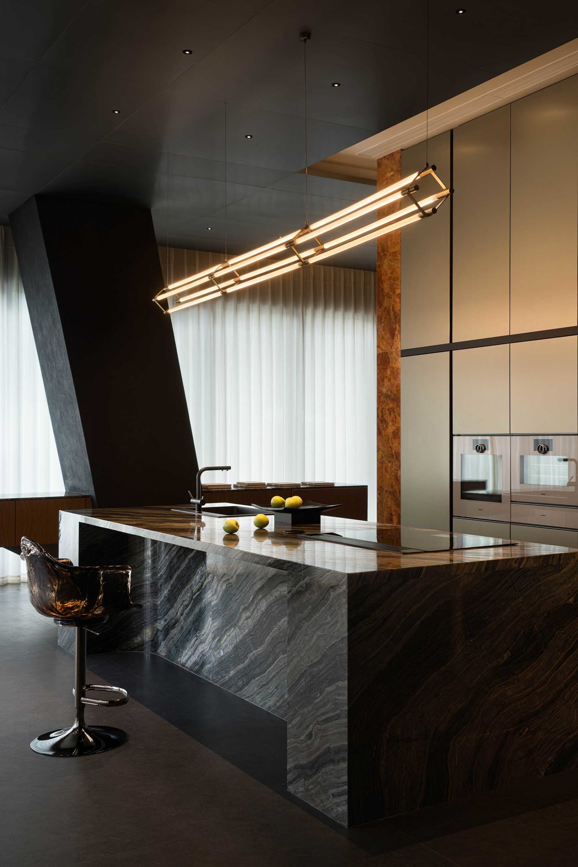 Modern luxury kitchen featuring a large dark marble island with integrated cooktop and sink, a transparent amber bar stool, and a long linear gold pendant light hanging above.