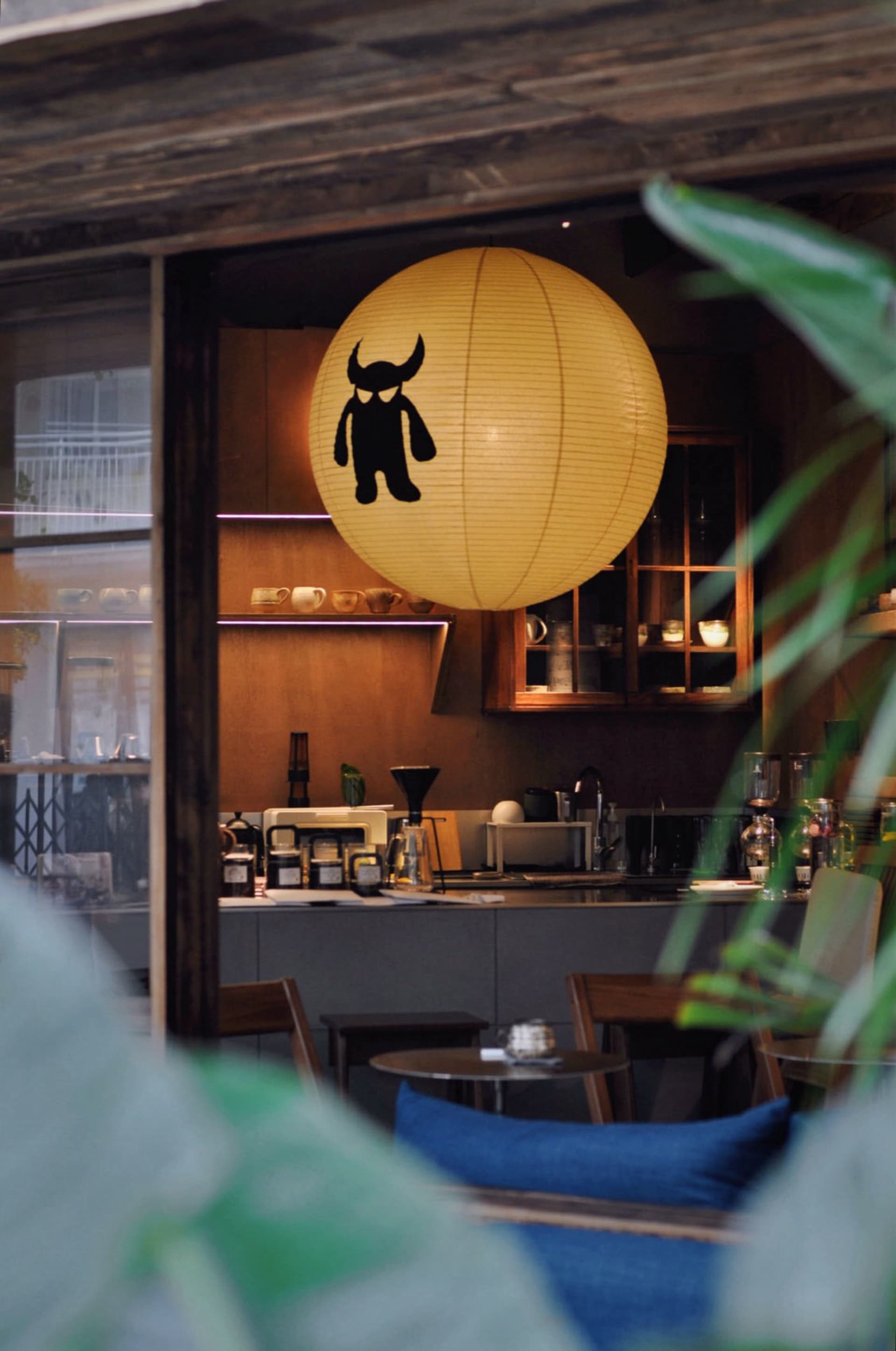 Interior view of a cozy cafe featuring a large yellow paper lantern with a black monster design hanging above a wooden coffee counter with warm lighting and wooden furniture.