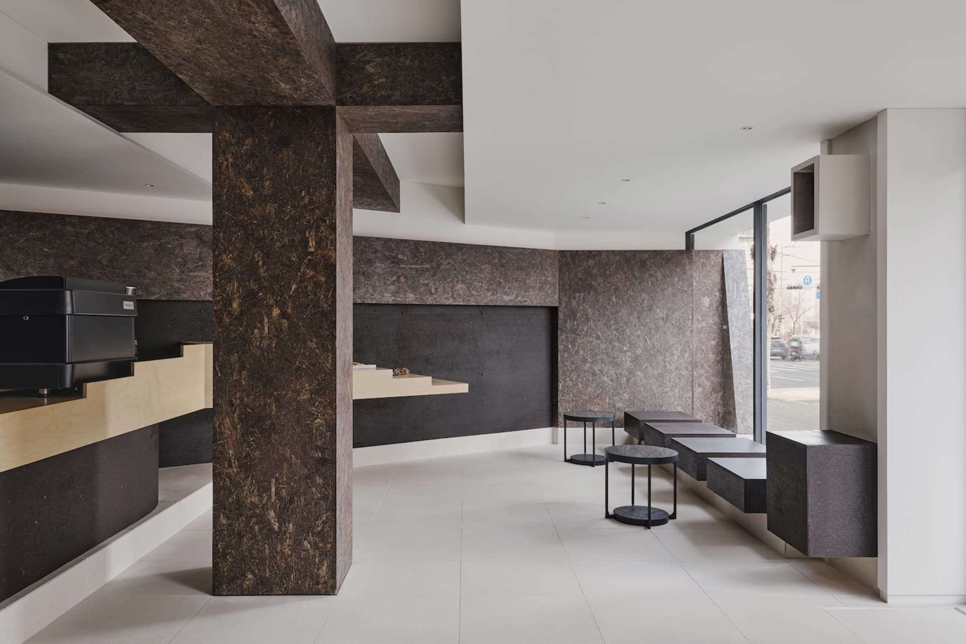 Modern minimalist cafe interior featuring dark marble walls, large structural beams, floating black stone benches and round side tables against a bright white floor.