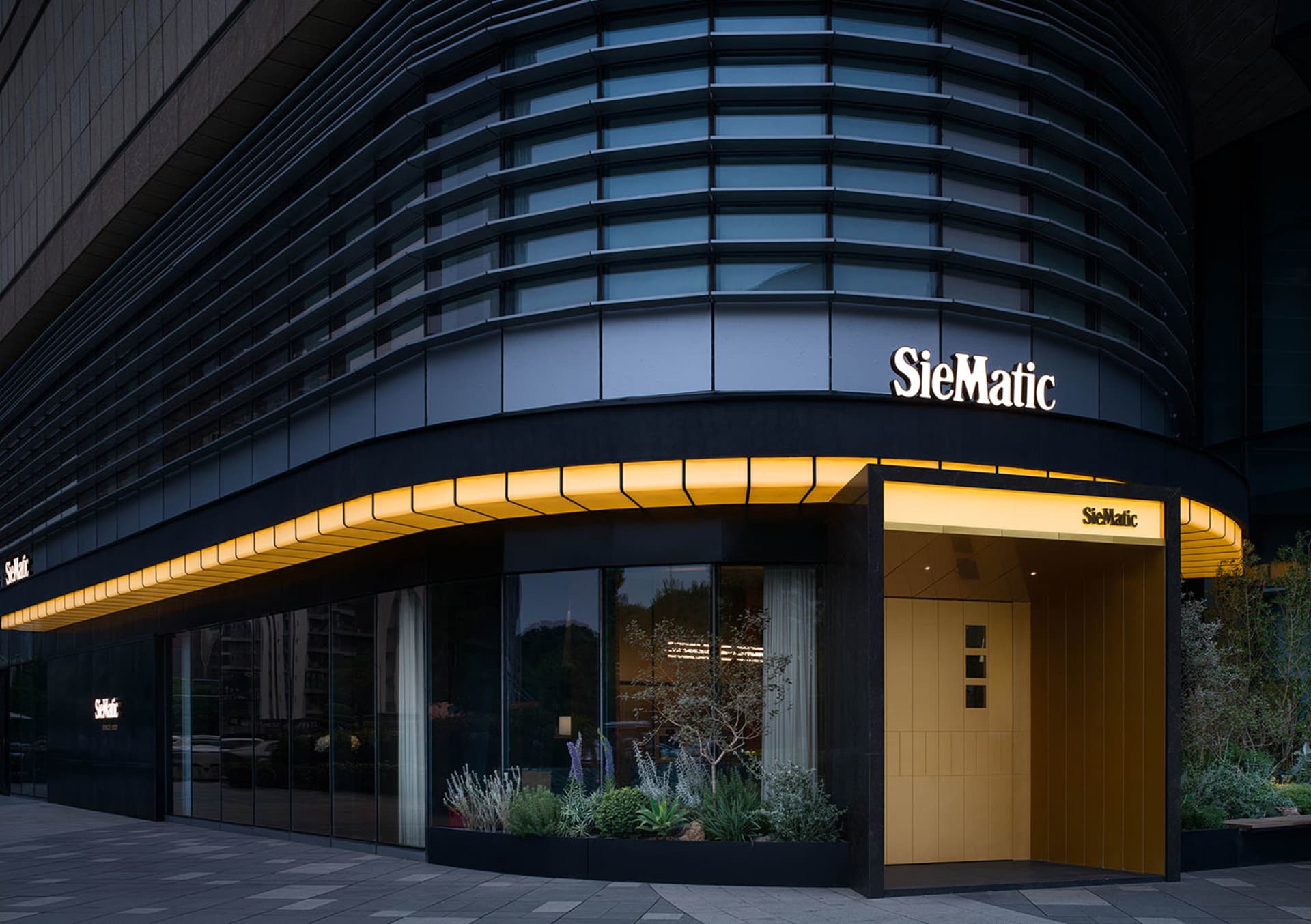 Night view of a modern luxury kitchen showroom with a curved dark glass facade, warm yellow LED lighting along the canopy, and landscaped planters at the entrance.