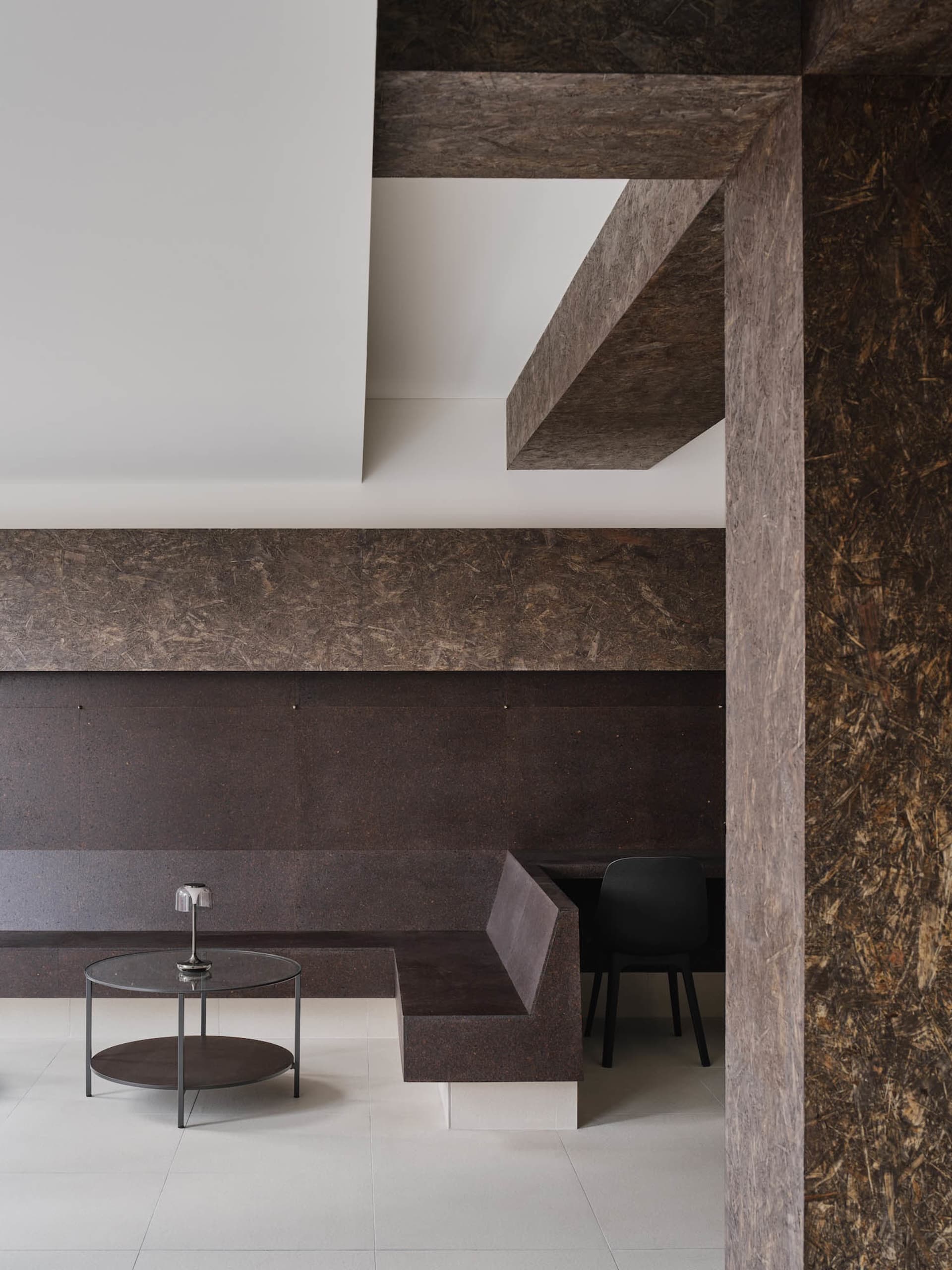 Modern minimalist entryway featuring a large raw OSB feature wall, built-in dark bench, round glass coffee table with lamp, and black chair.