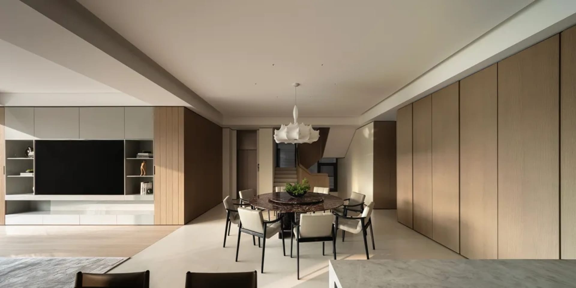 Modern minimalist dining room featuring a large round dark marble table surrounded by six light-colored dining chairs, under a white sculptural pendant lamp, with floor-to-ceiling walnut wood paneling and built-in media console in the background.