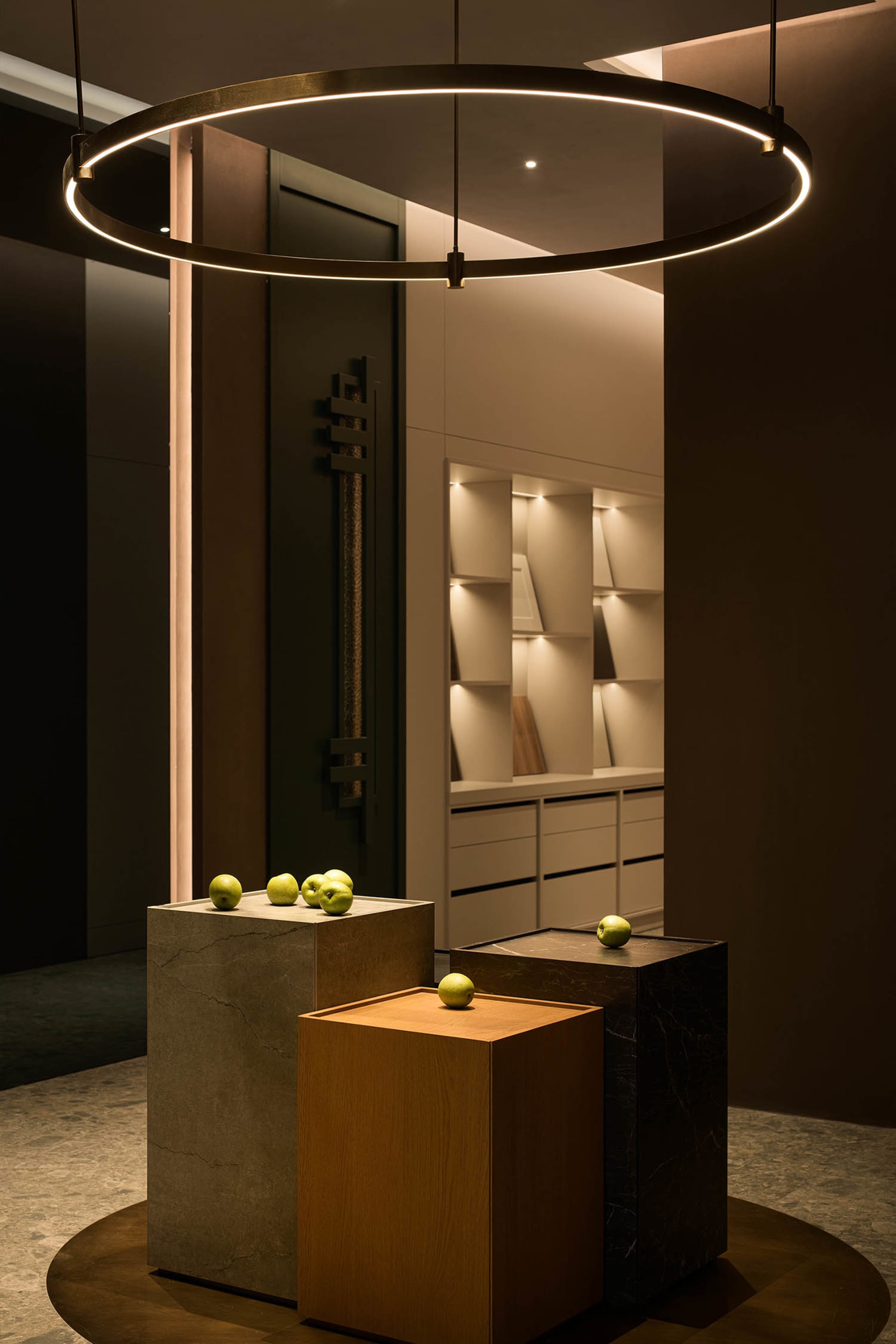 Modern minimalist entryway featuring a large circular LED pendant lamp, three display pedestals in concrete, light oak, and black marble, each topped with green apples, with built-in illuminated shelving in the background.