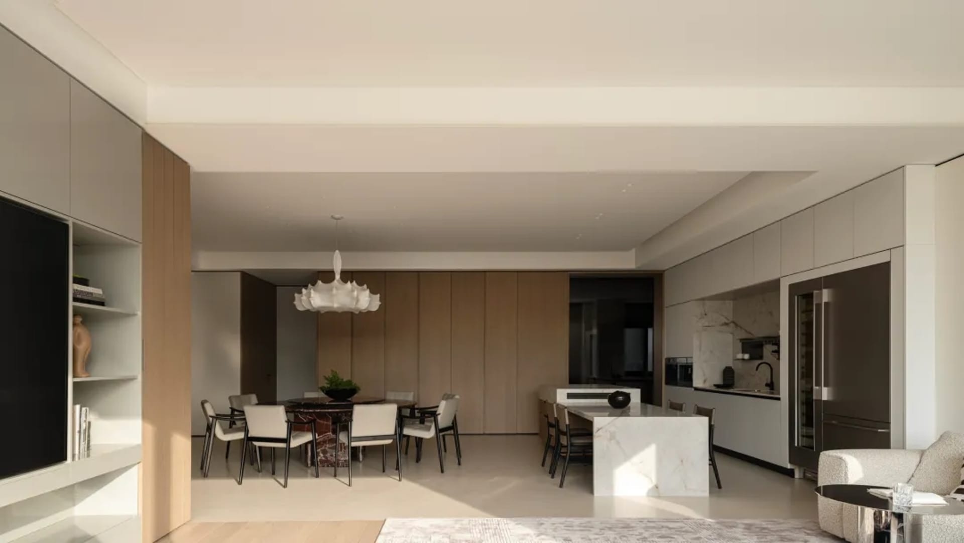 Modern open-plan apartment interior featuring a dining table with six chairs, a large white marble kitchen island, light wood cabinetry, and a sculptural white chandelier.