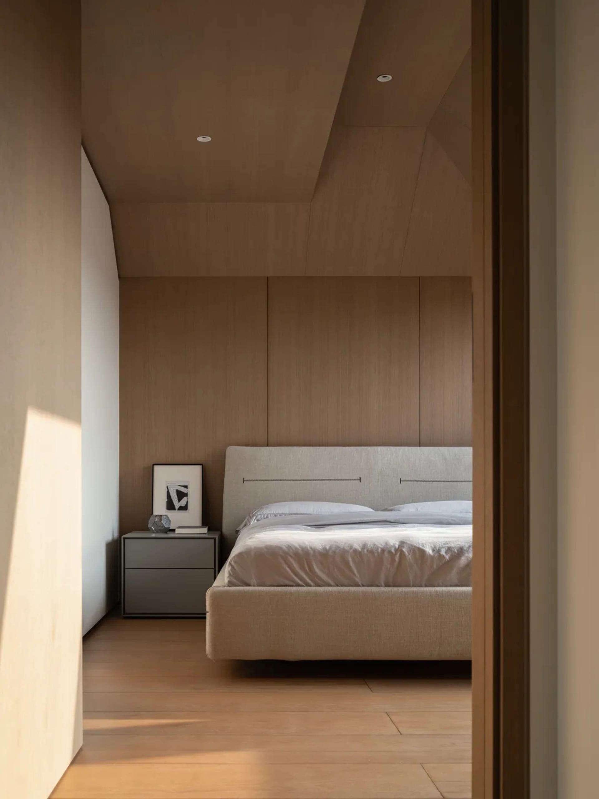View into a minimalist bedroom featuring a beige upholstered bed, wood-paneled walls and ceiling, and a grey floating nightstand with framed art.