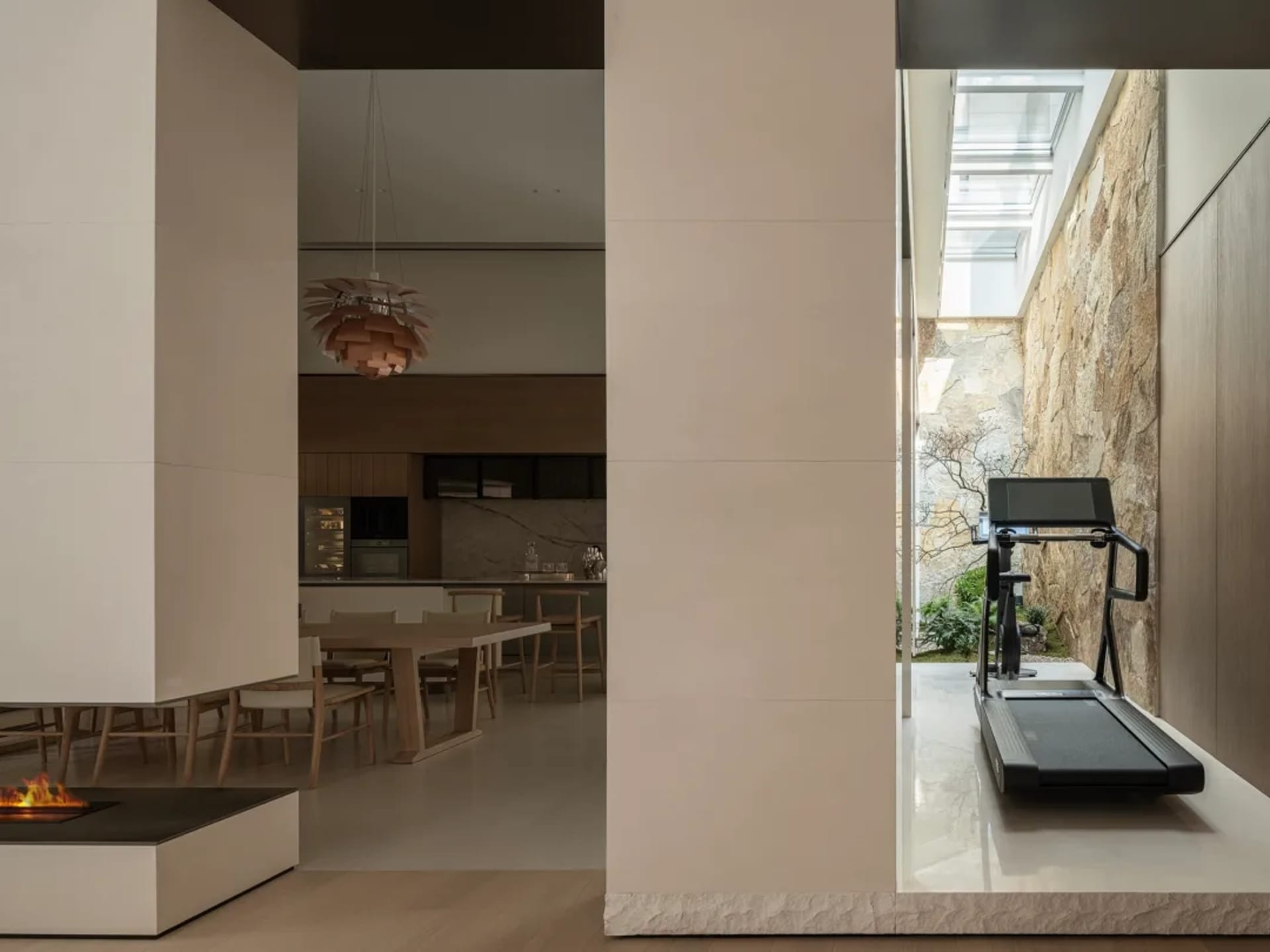 Modern open-plan interior showing a dining area, kitchen, fireplace, wooden pendant light, and a treadmill facing a stone-walled courtyard with skylight.