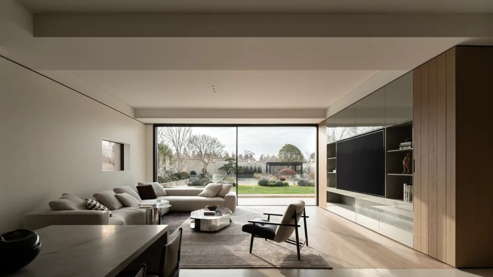Modern minimalist living room featuring a large L-shaped sectional sofa, oak wood entertainment wall with built-in TV and shelving, marble coffee table, black leather armchair, and floor-to-ceiling glass doors opening to a landscaped garden.