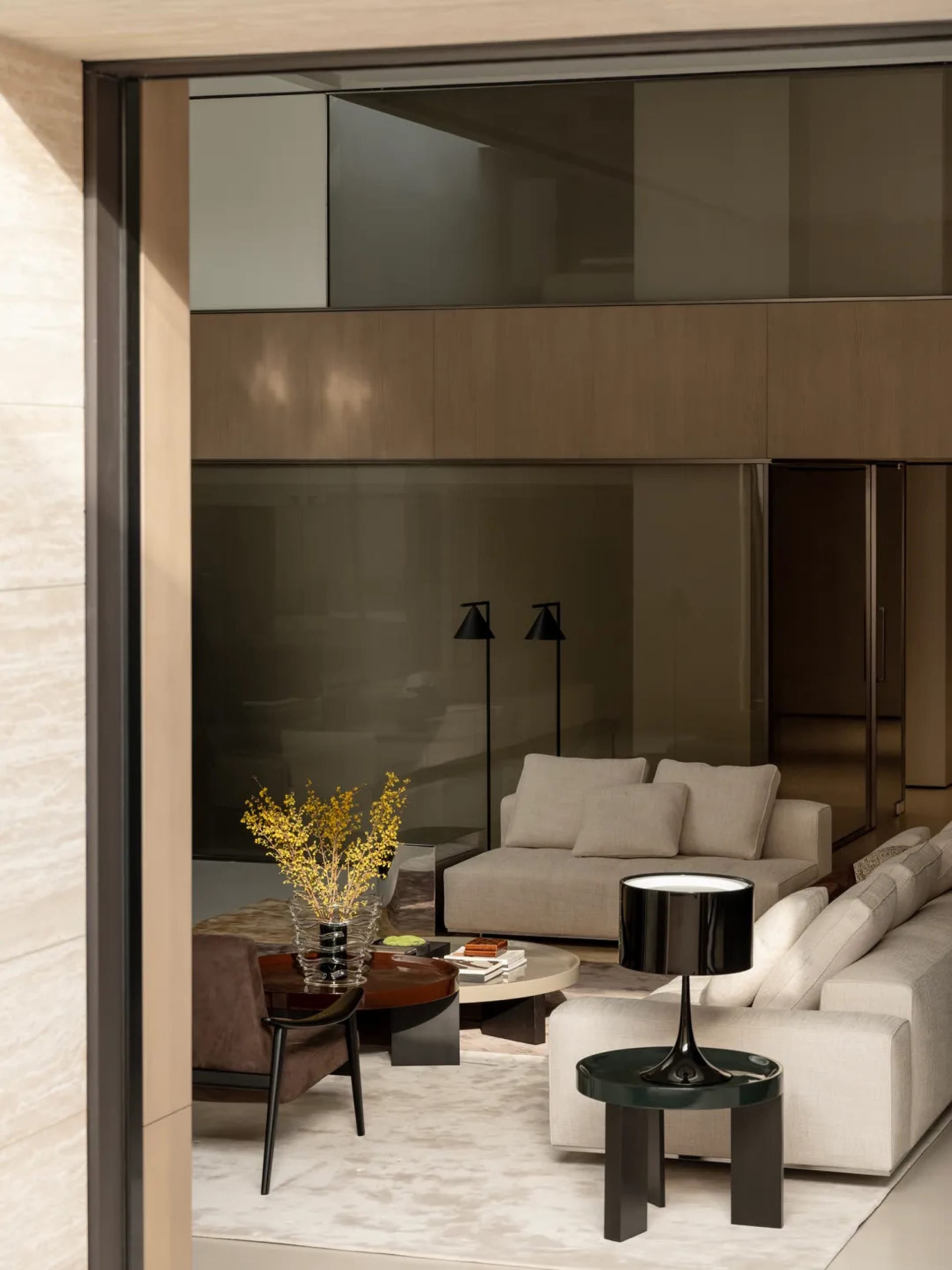 Modern luxury living room featuring a large beige sectional sofa, matching armchair, round coffee tables, black floor lamps, and warm wood paneling in the background.