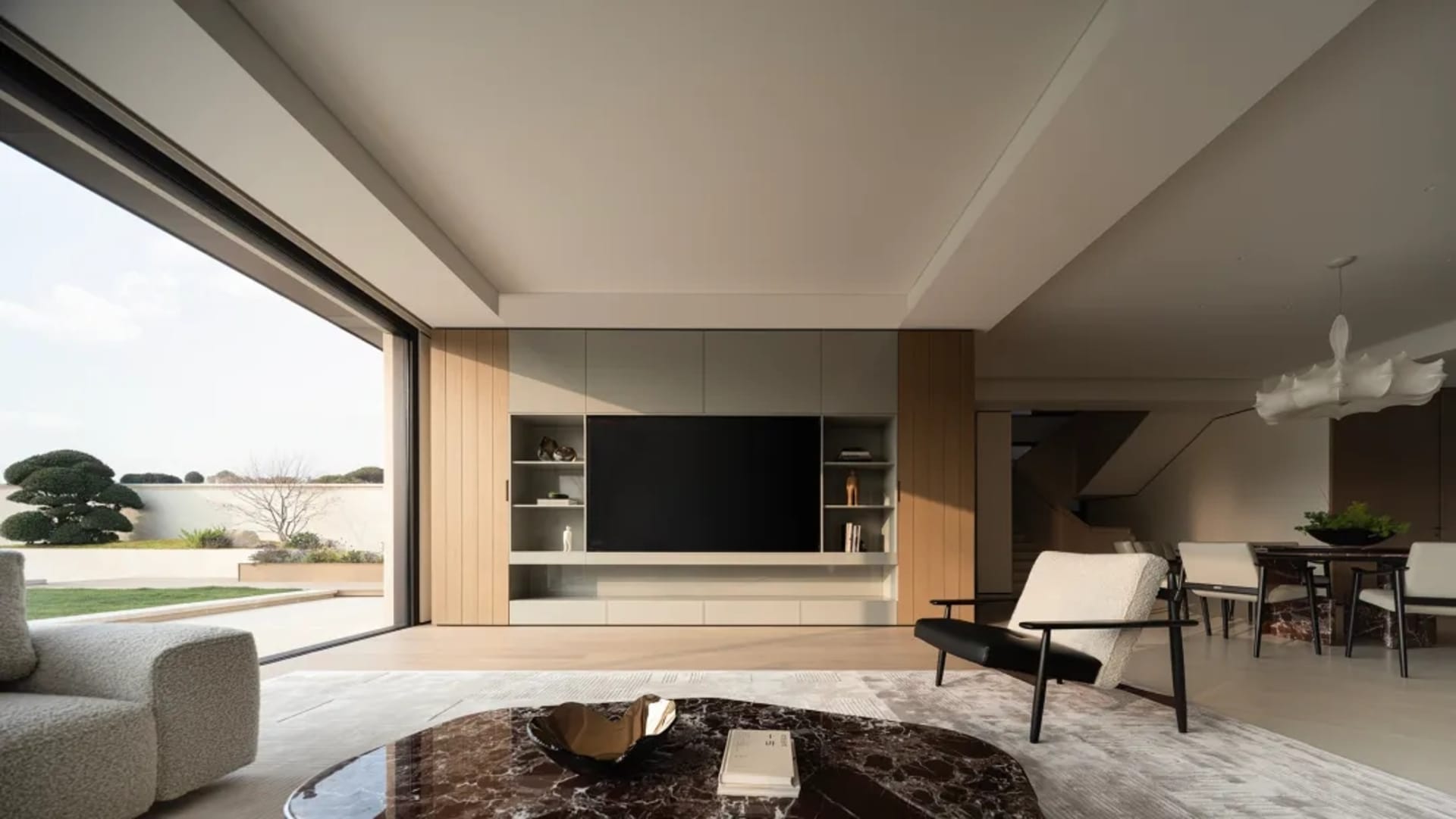 Modern minimalist living room featuring a walnut entertainment center with built-in TV, dark marble coffee table, light grey sofa, and dining area in the background with a sculptural white chandelier.