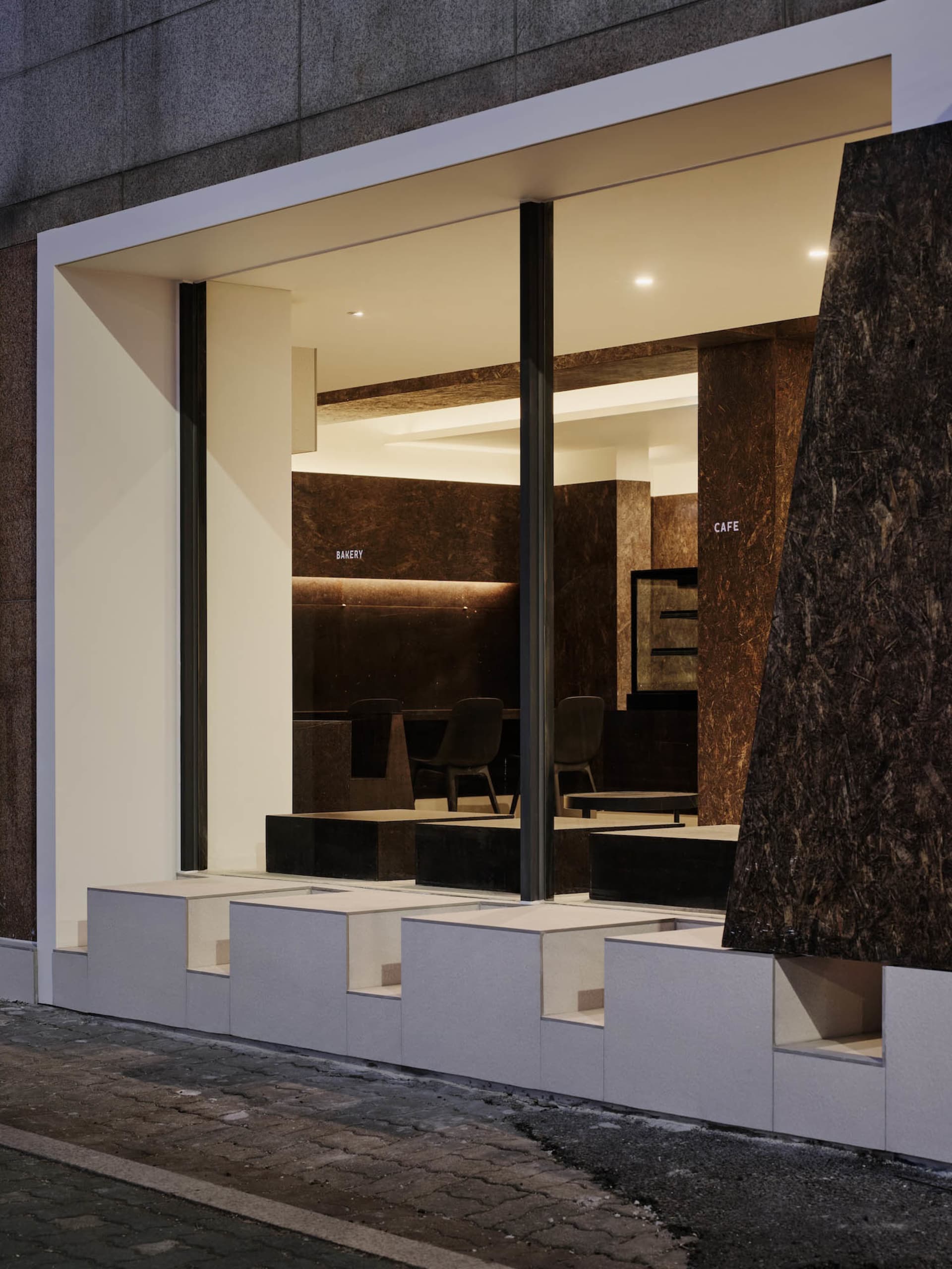 Night view of a modern minimalist cafe with white marble stepped seating and dark brown marble interior walls visible through large glass windows.