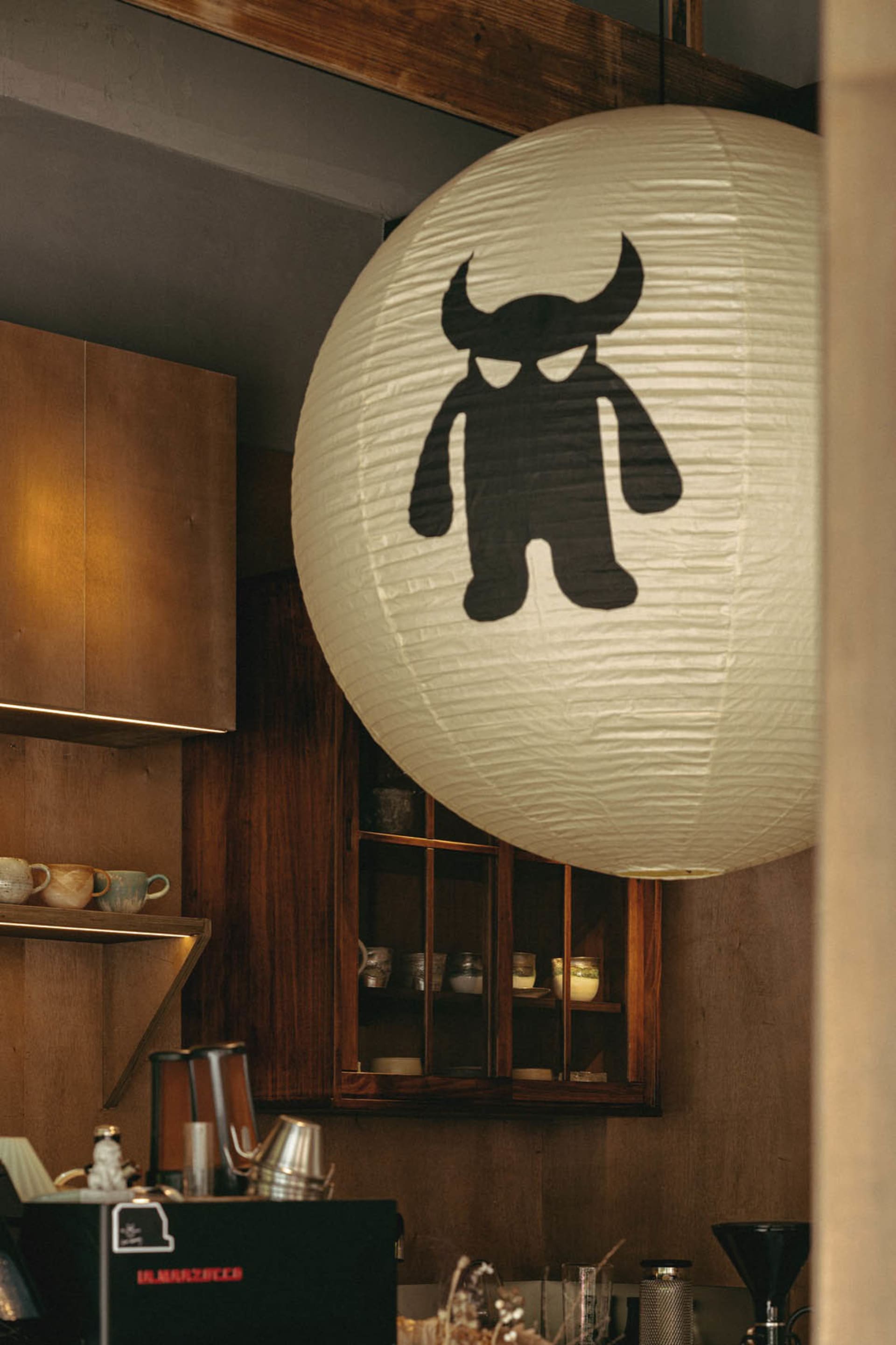 Interior view of a specialty coffee bar featuring warm wooden cabinetry, open shelving with ceramic cups, and a large white paper lantern with a black monster illustration hanging prominently in the foreground.