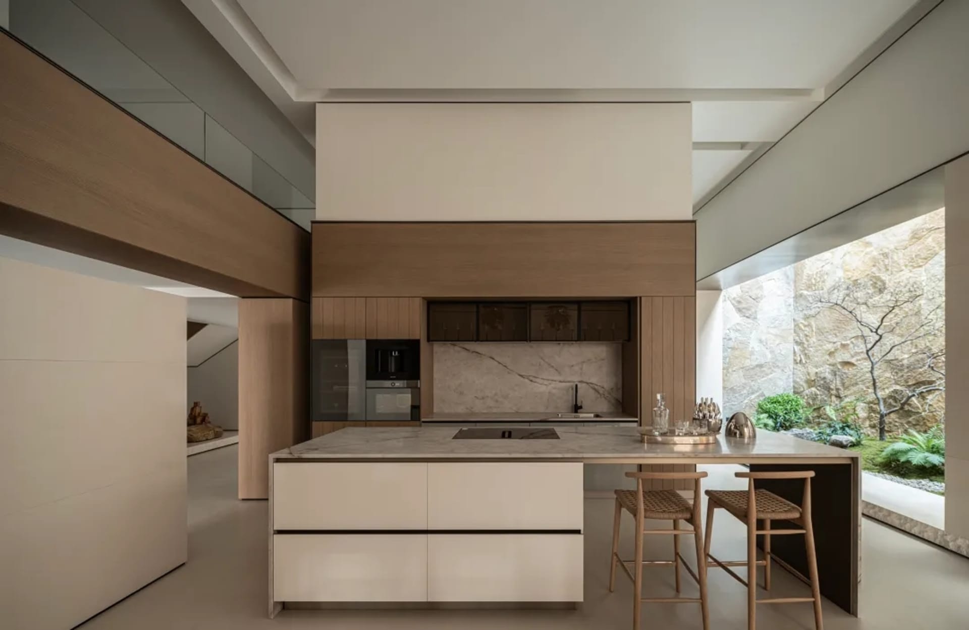 Modern minimalist kitchen featuring a large marble island with wooden bar stools, integrated wood cabinetry, and a large window overlooking a rock garden.