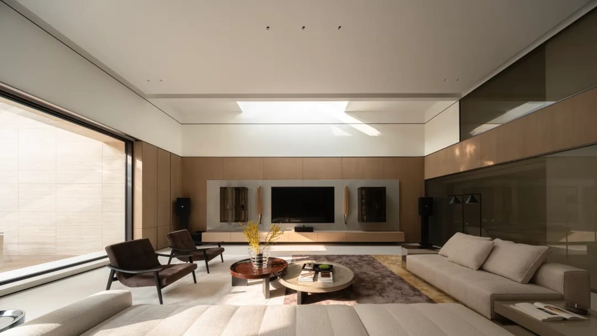 Modern minimalist living room with beige sofa, dark brown leather armchairs, round coffee tables on a rug, and a large walnut entertainment center with wall-mounted TV under a skylight.