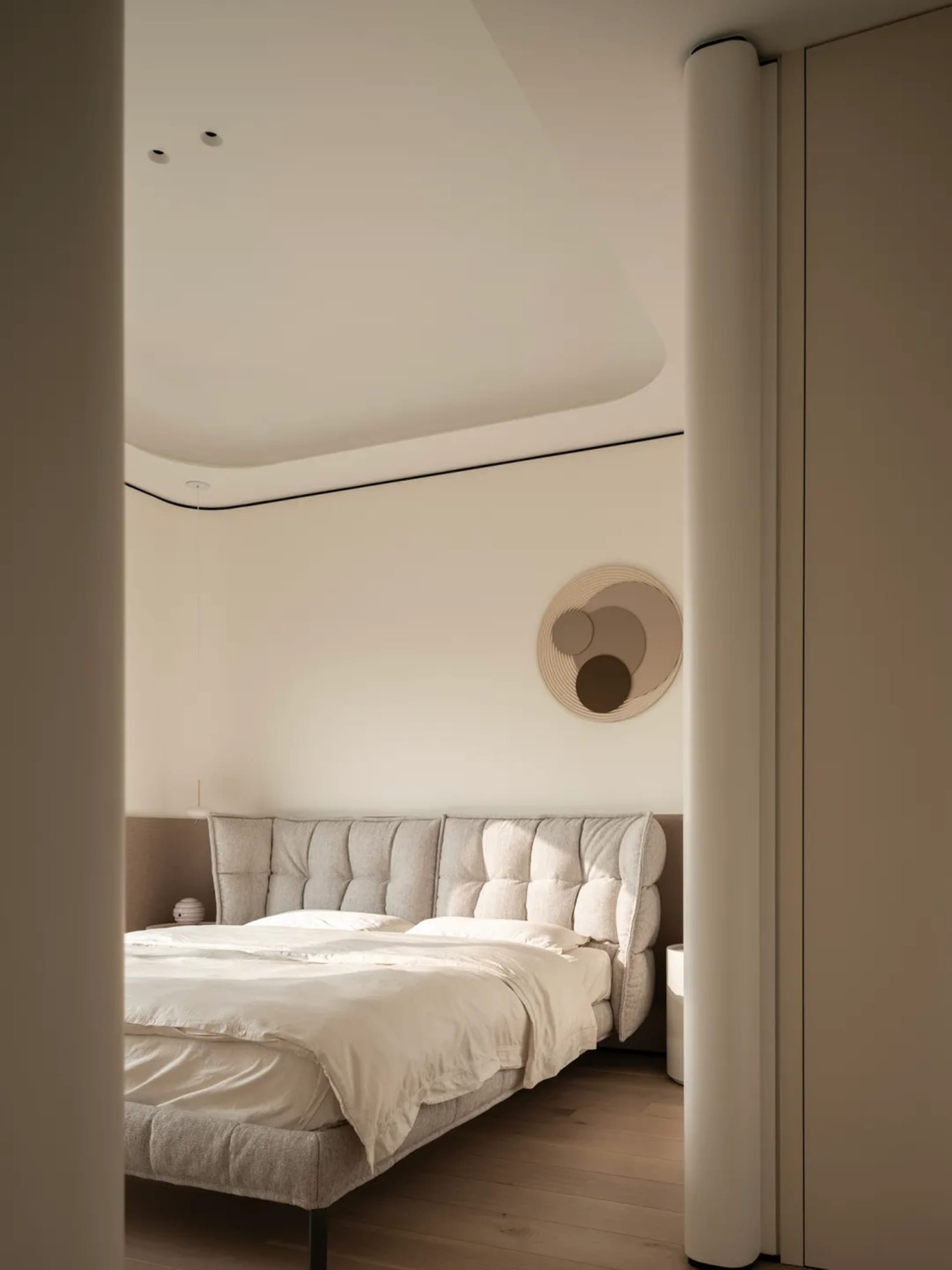 Modern minimalist bedroom with a large tufted boucle fabric bed, white bedding, round woven wall art, and curved ceiling details in neutral tones.