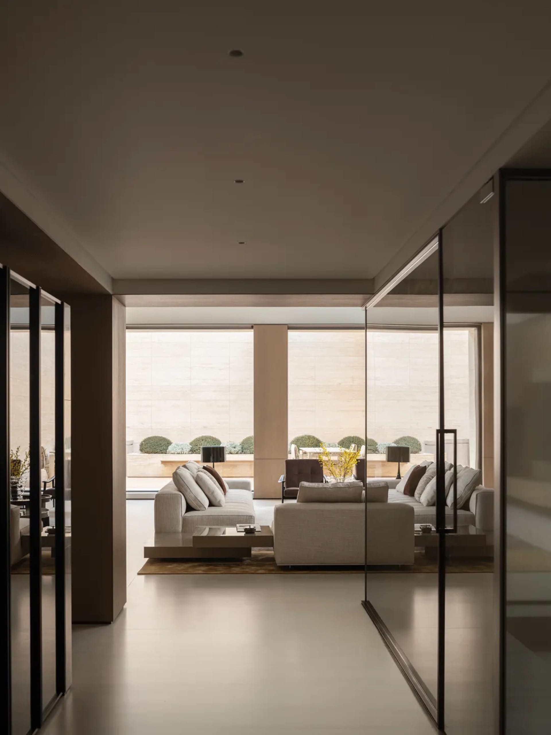Modern minimalist living room featuring light travertine walls and floor, neutral fabric sofas, and large glass doors opening to a landscaped garden with trimmed shrubs.