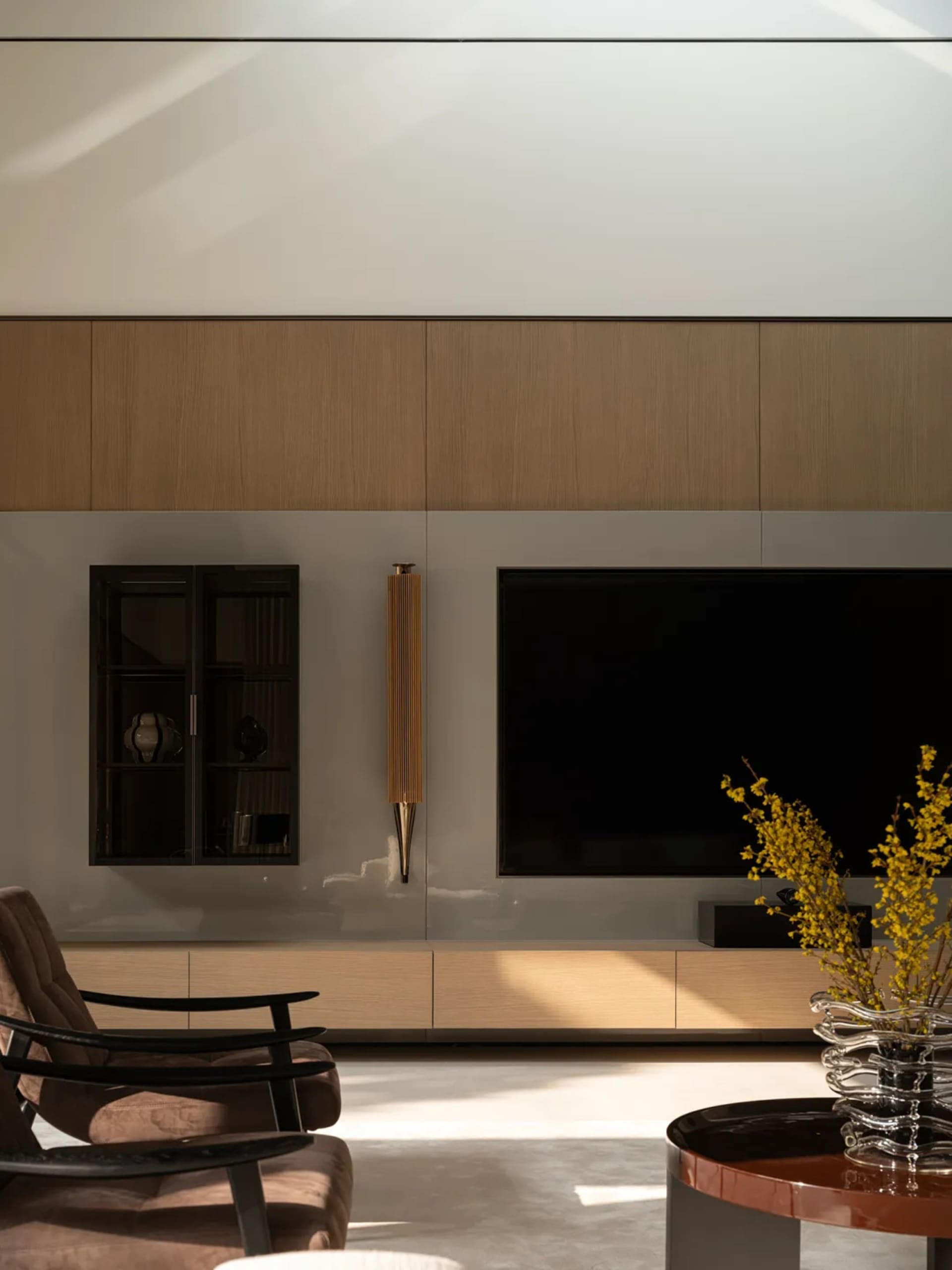 Modern minimalist living room featuring a large oak wood panel entertainment wall, wall-mounted TV, dark wood display cabinet, brown upholstered armchair with black frame, and a glossy round coffee table with yellow forsythia branches.
