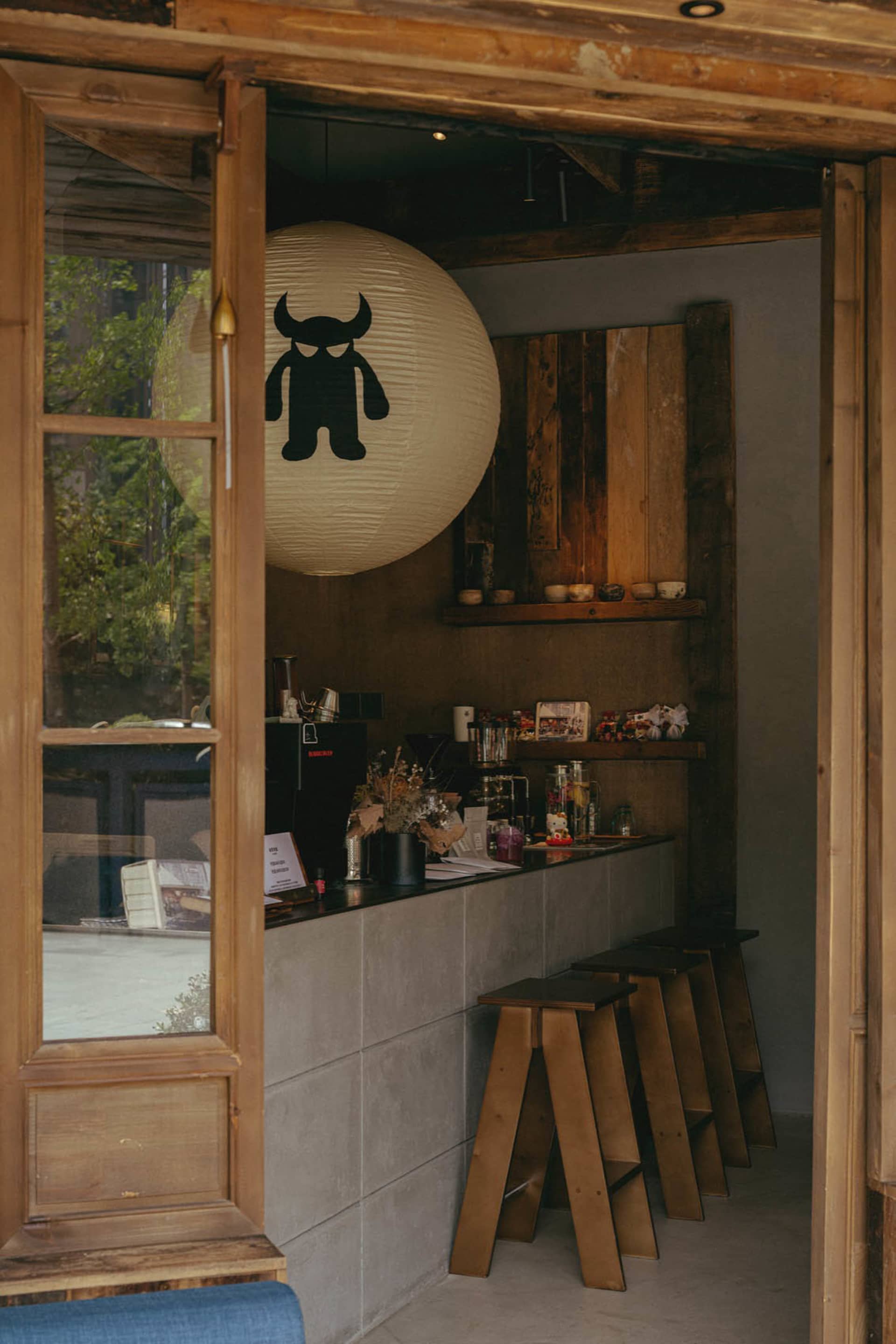 View through a wooden doorway into a small cafe featuring a concrete counter, wooden stools, a large white paper lantern with a black monster graphic, and rustic wooden wall paneling.