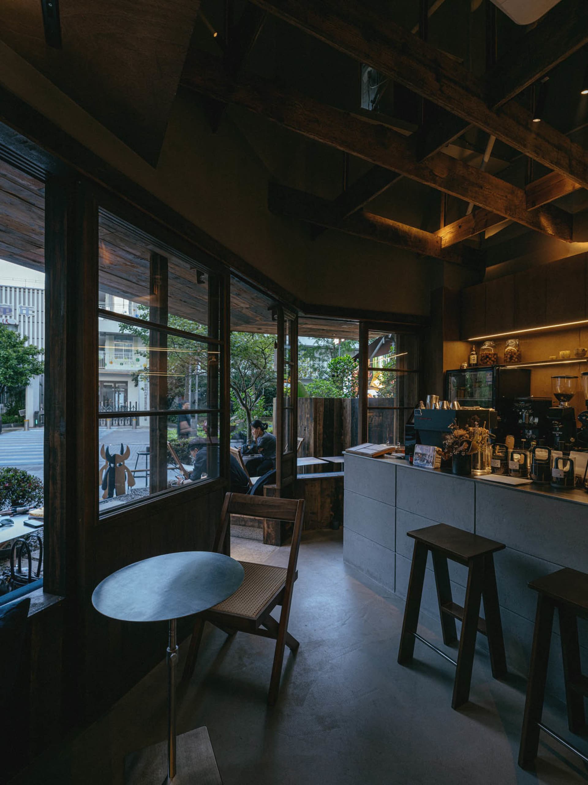 Interior view of a modern Taipei cafe featuring dark reclaimed wood ceiling beams, a concrete service counter, wooden stools, and large glass windows overlooking a tree-lined street.