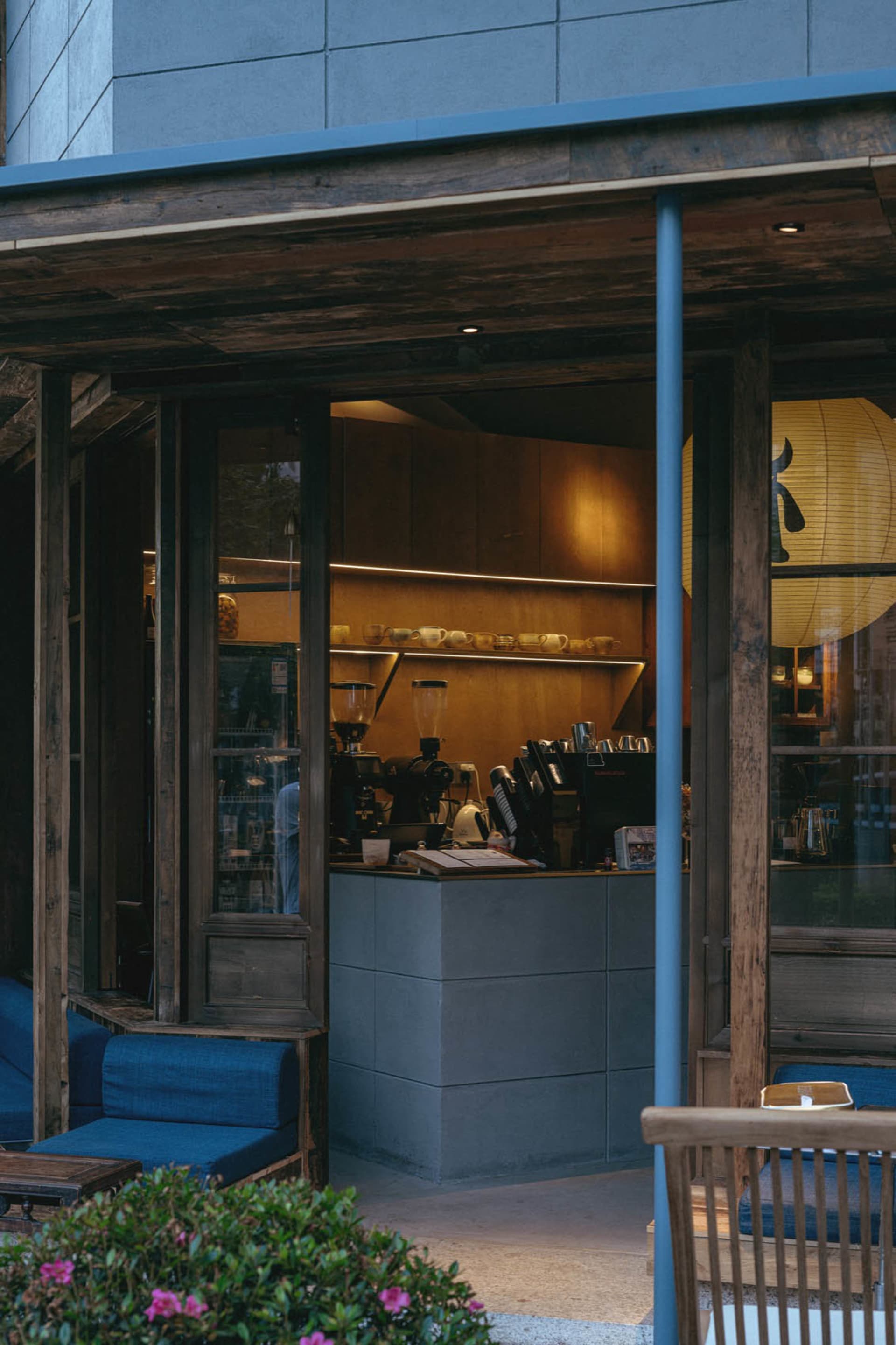 View from an outdoor wooden terrace into a warmly lit Japanese-style cafe interior featuring concrete counter, reclaimed wood beams, and deep blue upholstered seating.