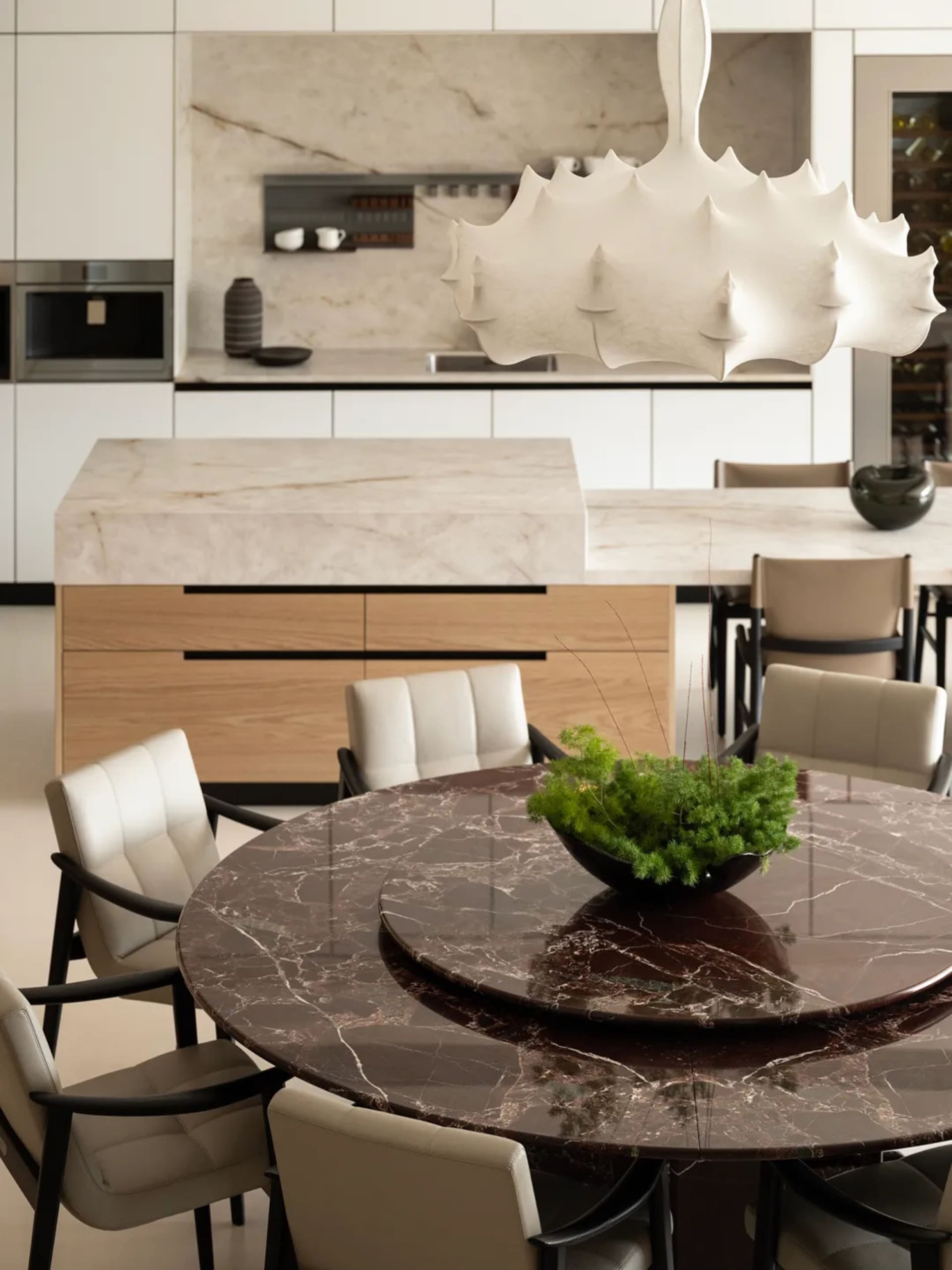 Modern dining area featuring a large round dark brown marble table surrounded by cream leather dining chairs, with a sculptural white pendant lamp hanging above and an open kitchen with light marble island in the background.