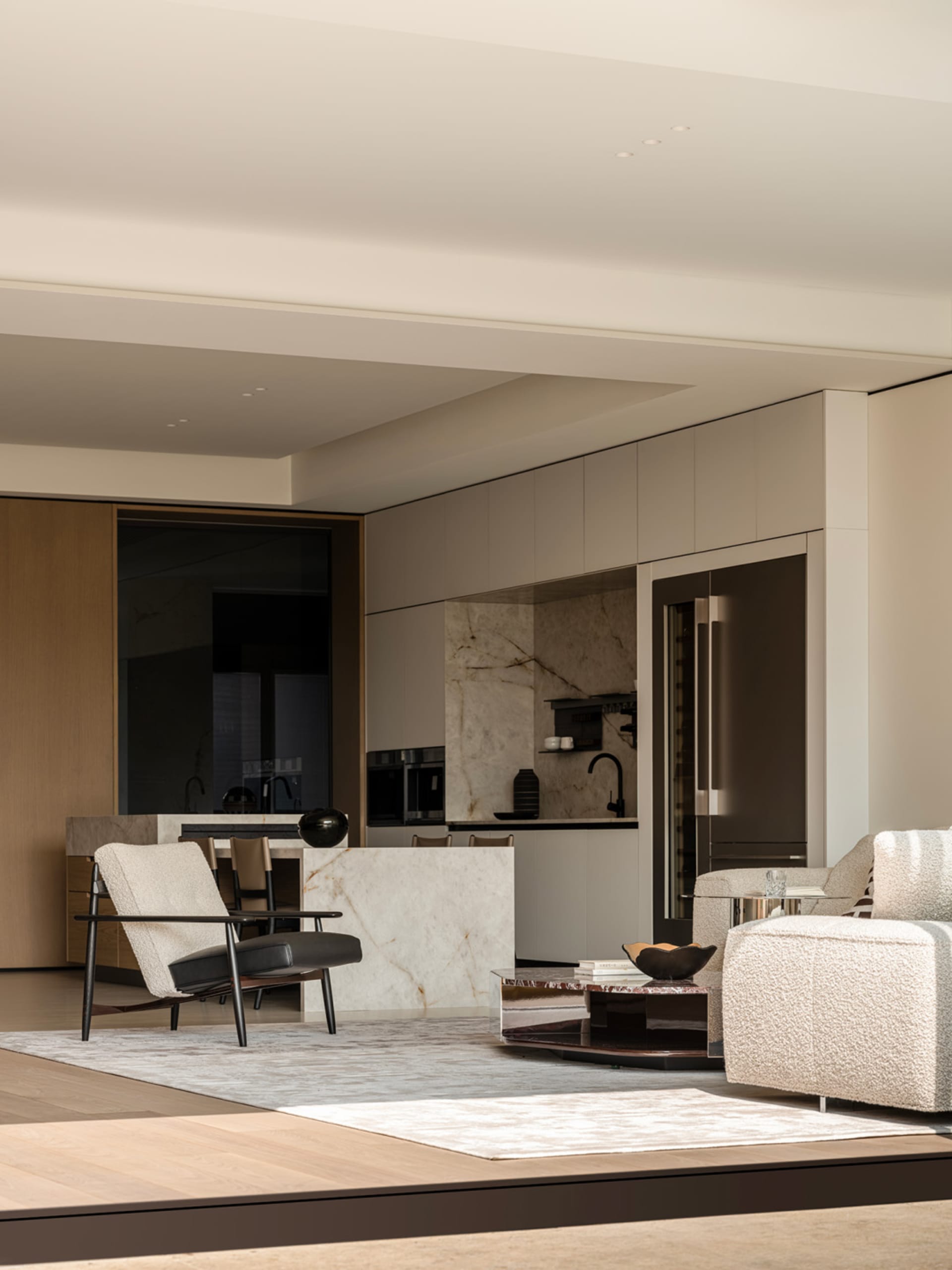 Modern minimalist living room featuring a white marble kitchen island, boucle sofa, black leather and wood armchair, and integrated stainless steel appliances.
