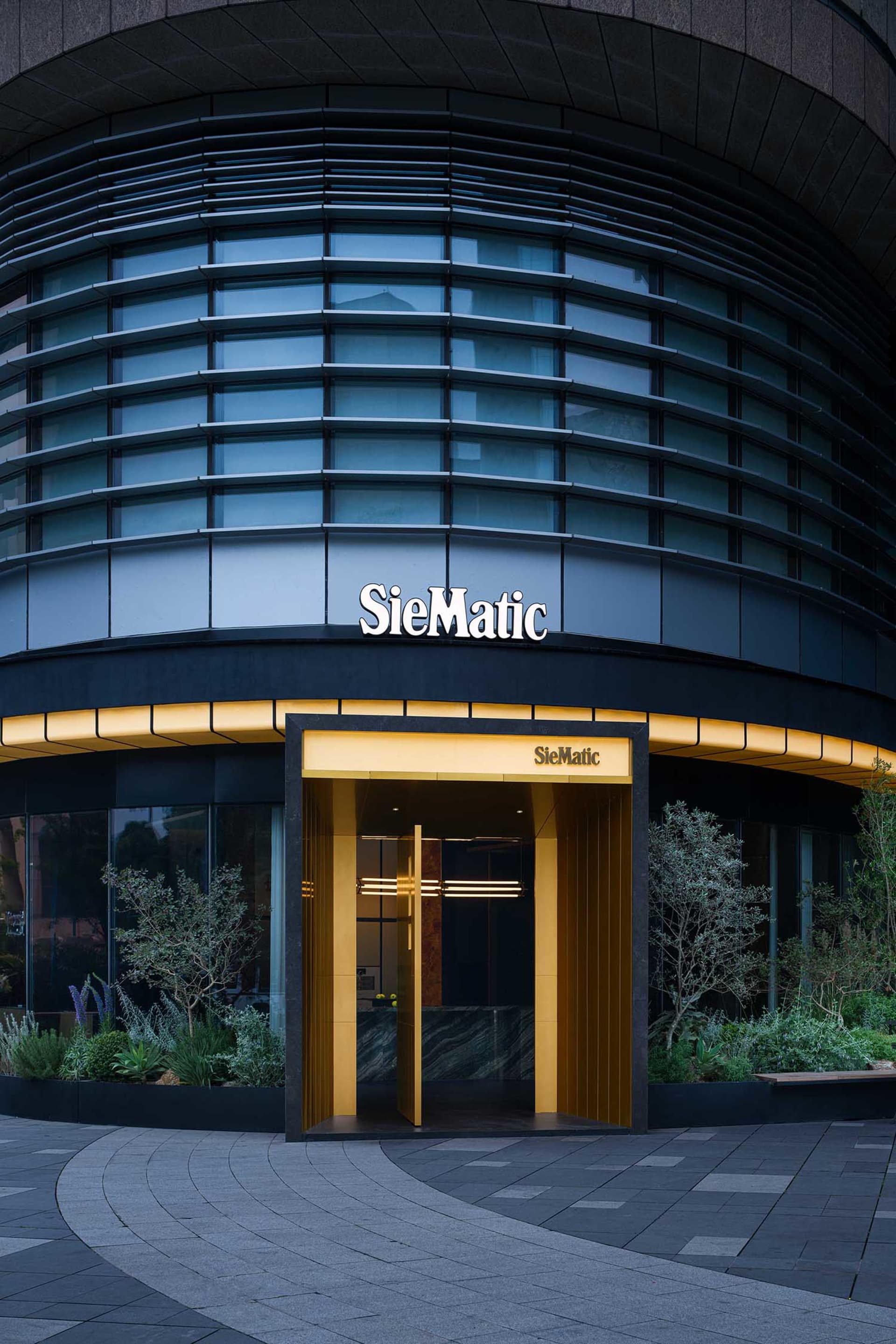 Modern curved glass commercial building facade at night with illuminated golden entrance and SieMatic branding, surrounded by landscaped planters.