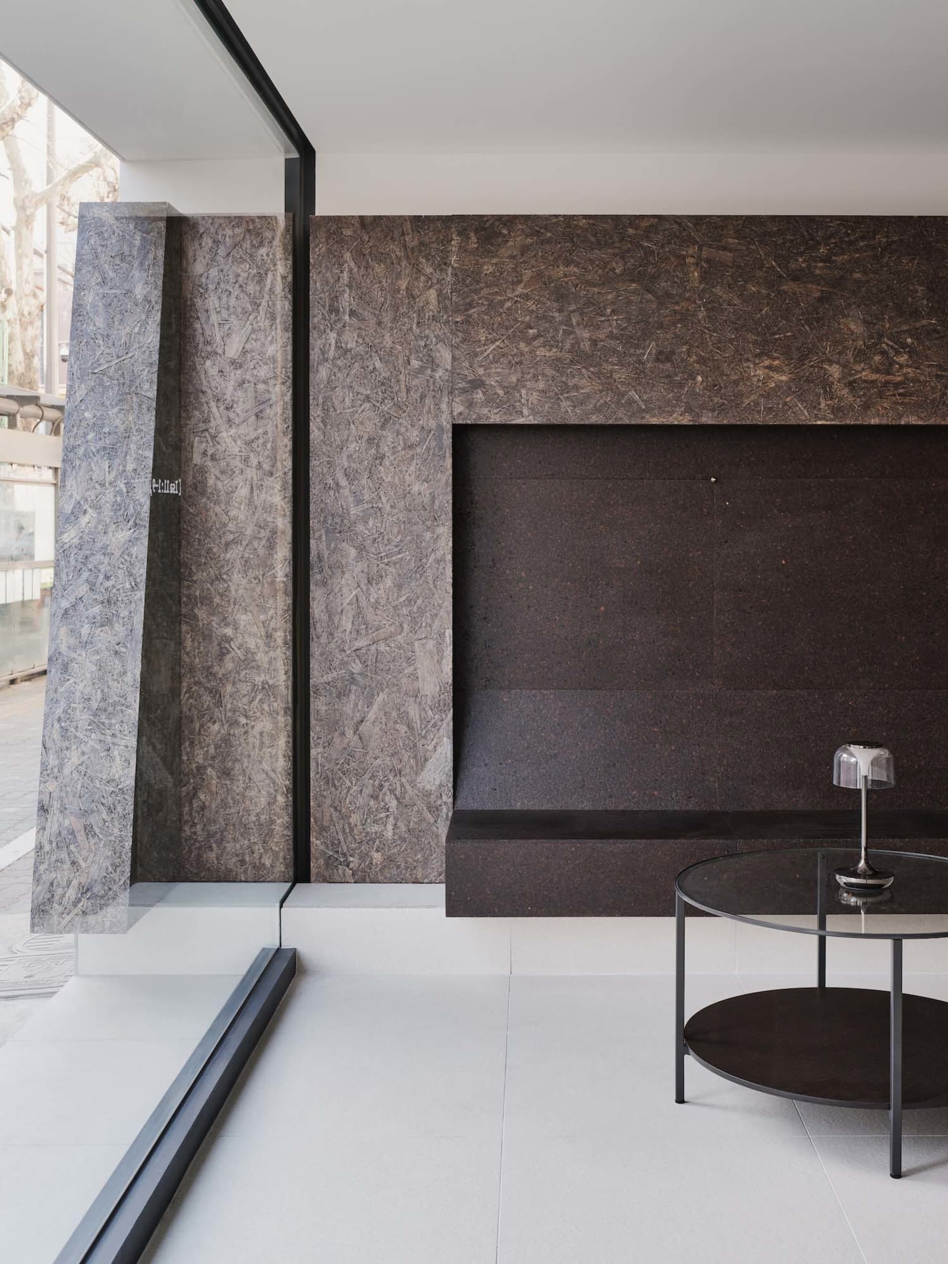 Modern minimalist living room featuring a large raw oriented strand board feature wall, concrete-like pillar, round glass coffee table, and a modern table lamp.