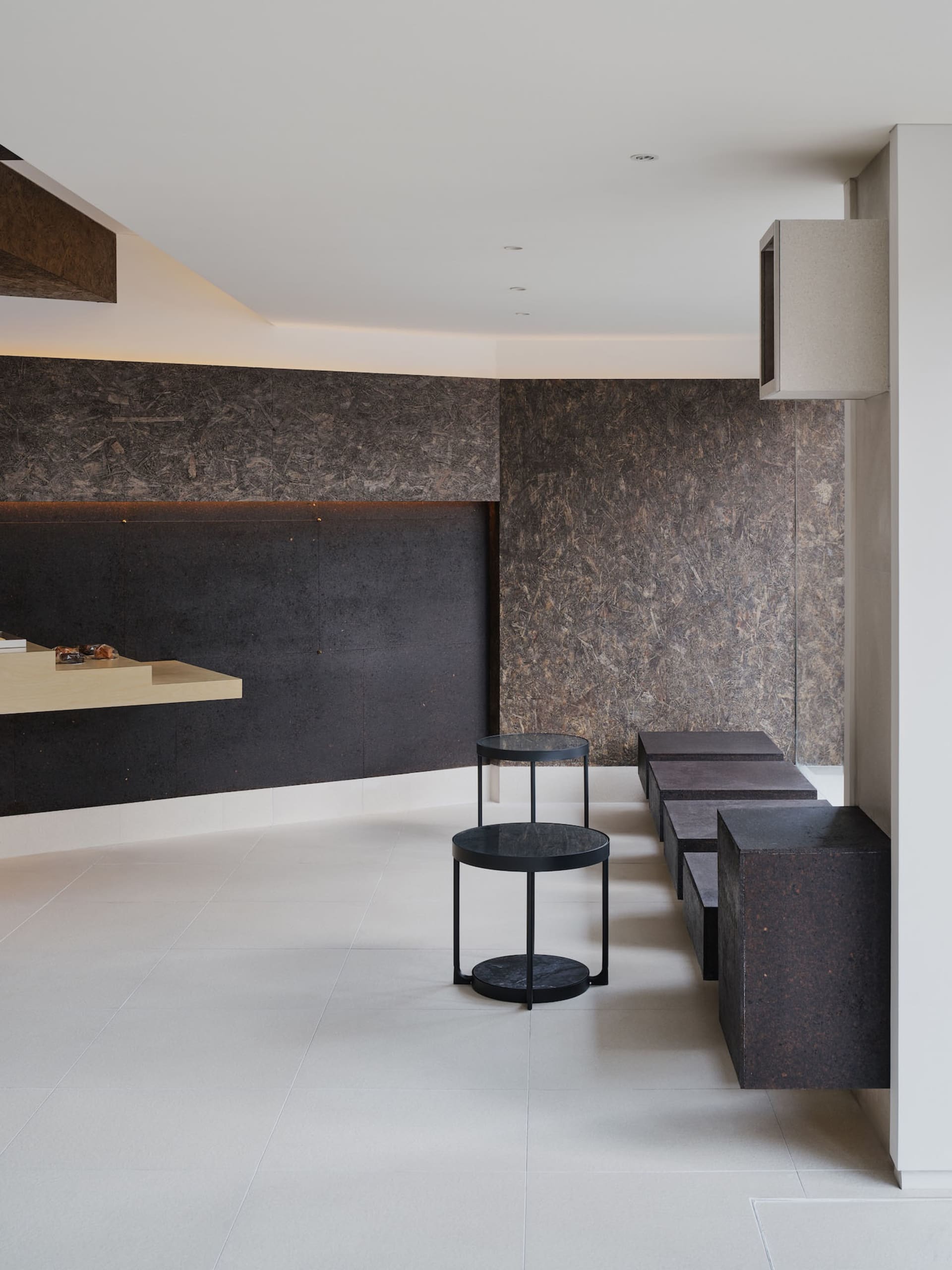 Modern minimalist living room featuring dark brown terrazzo accent wall and built-in bench, light wood floating console, black metal and terrazzo nesting tables, and white tiled floor.