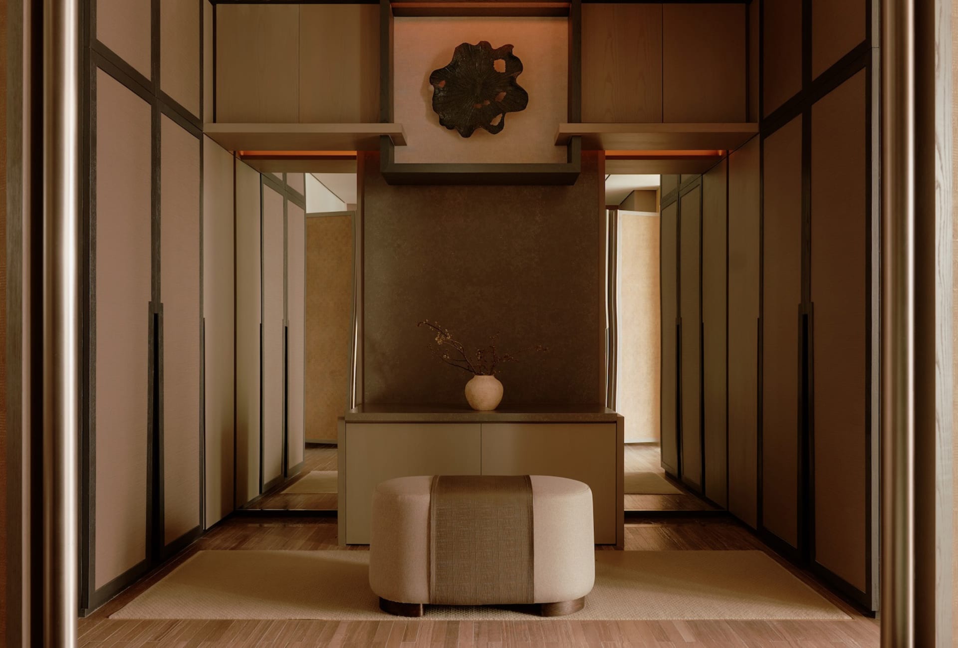 Modern minimalist entryway featuring a walnut console cabinet, upholstered ottoman with woven center band, dark metal leaf wall art, and warm ambient lighting in a neutral palette.
