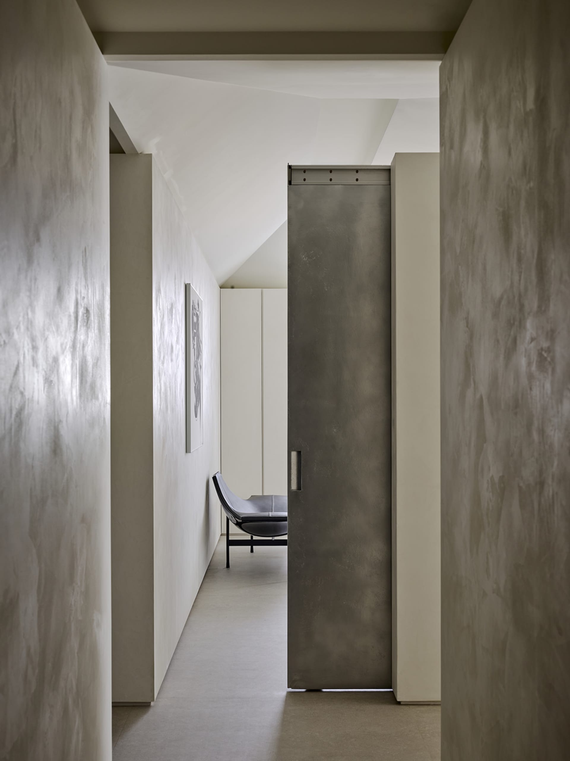 Narrow minimalist hallway with textured concrete walls, a large grey pivot door, and a black leather chaise lounge visible in the background under a sloped ceiling.