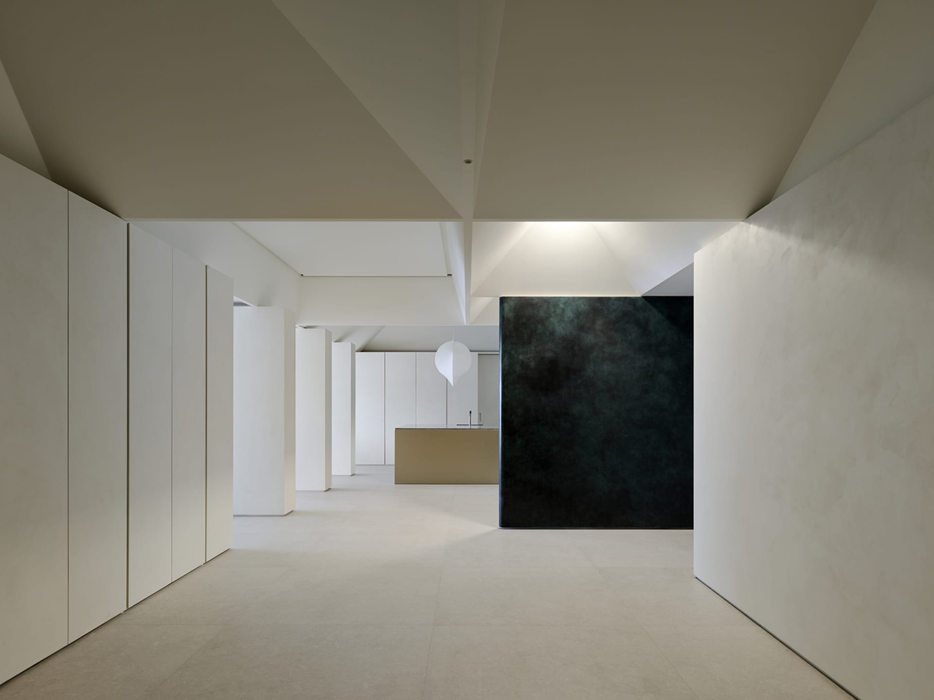 Modern minimalist interior hallway with white micro-cement walls and floor, leading to a kitchen island, featuring a large dark textured accent wall and a white pendant lamp.