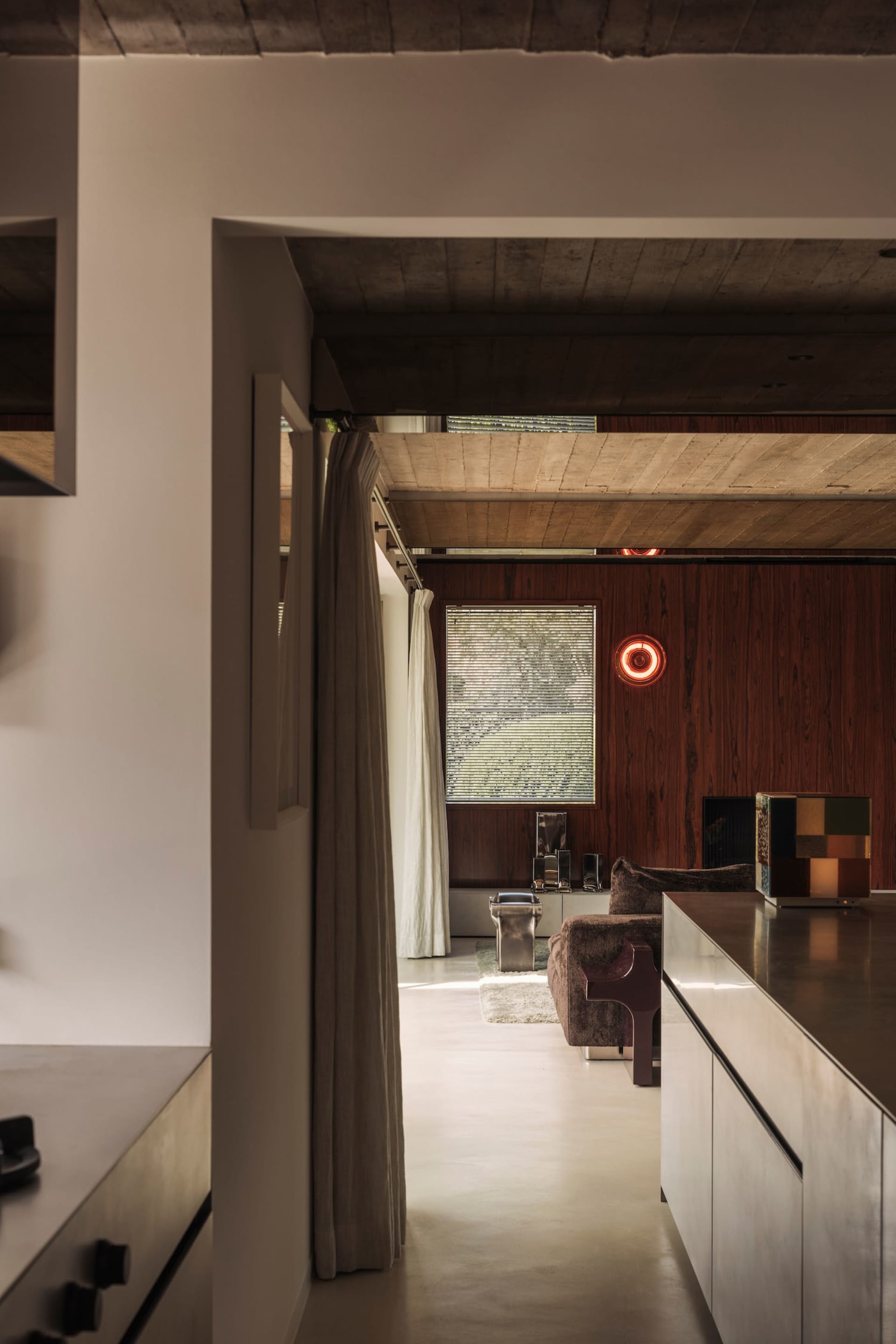 Interior view of a modern living room featuring a dark walnut wood accent wall, exposed concrete ceiling, brown bouclé sofa, and a glowing red circular wall light.