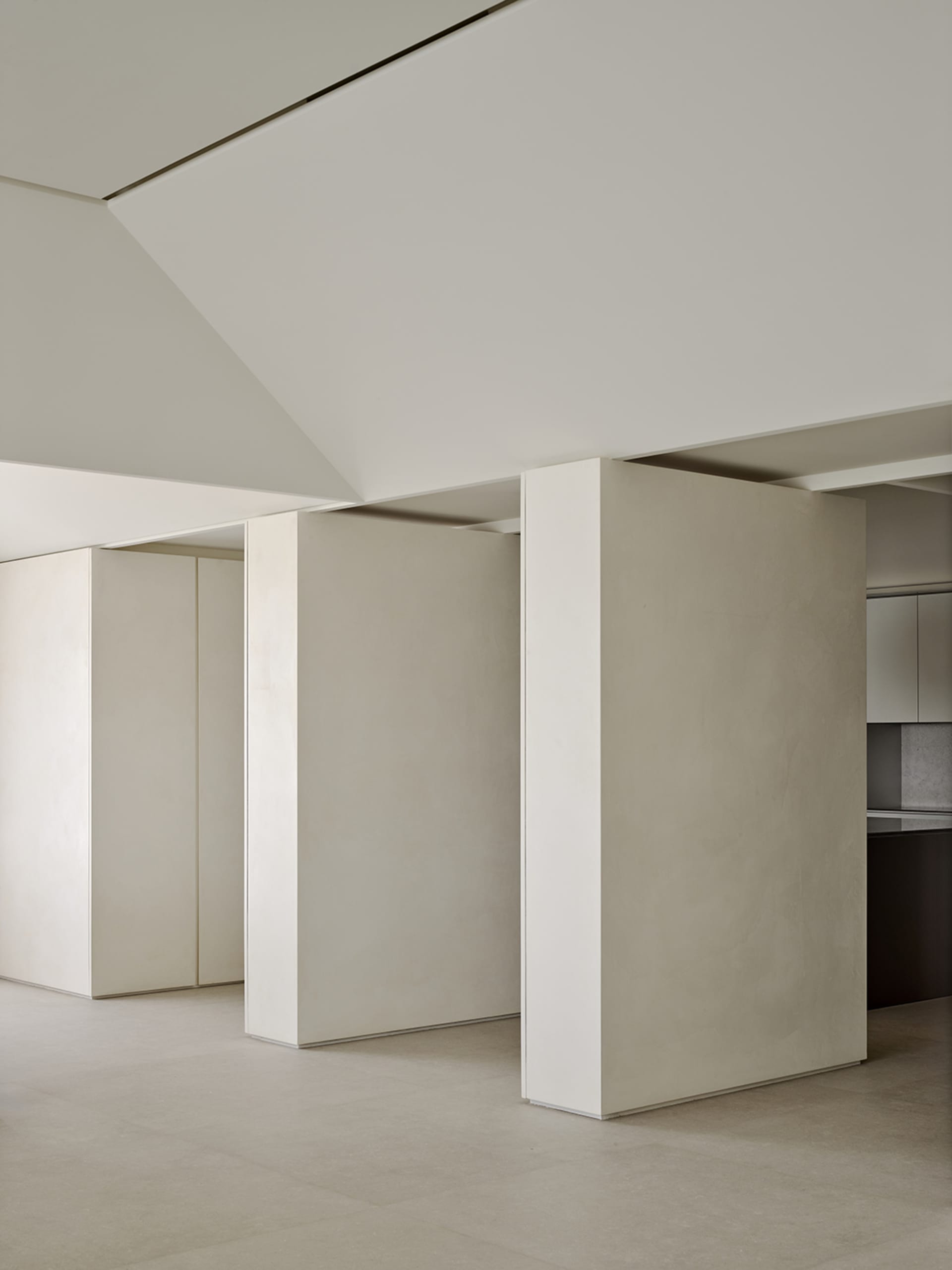 Minimalist living room interior featuring off-white micro-cement walls, a floating partition wall, high ceilings with recessed lighting, and seamless concrete flooring.