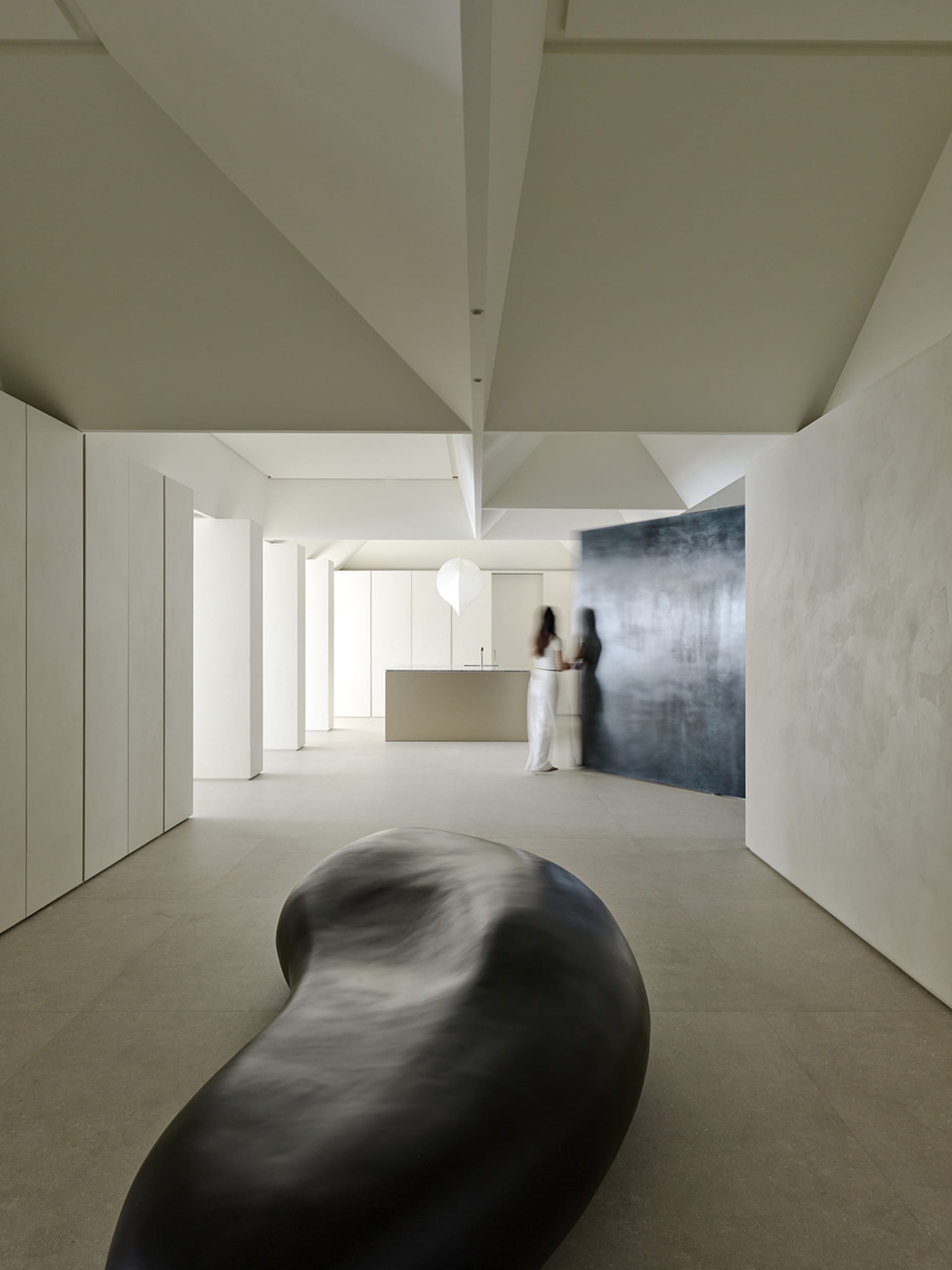 Minimalist interior hall featuring a large organic black sculptural bench in the foreground, textured micro-cement walls, high geometric ceiling, and a woman standing near a kitchen island in the background.