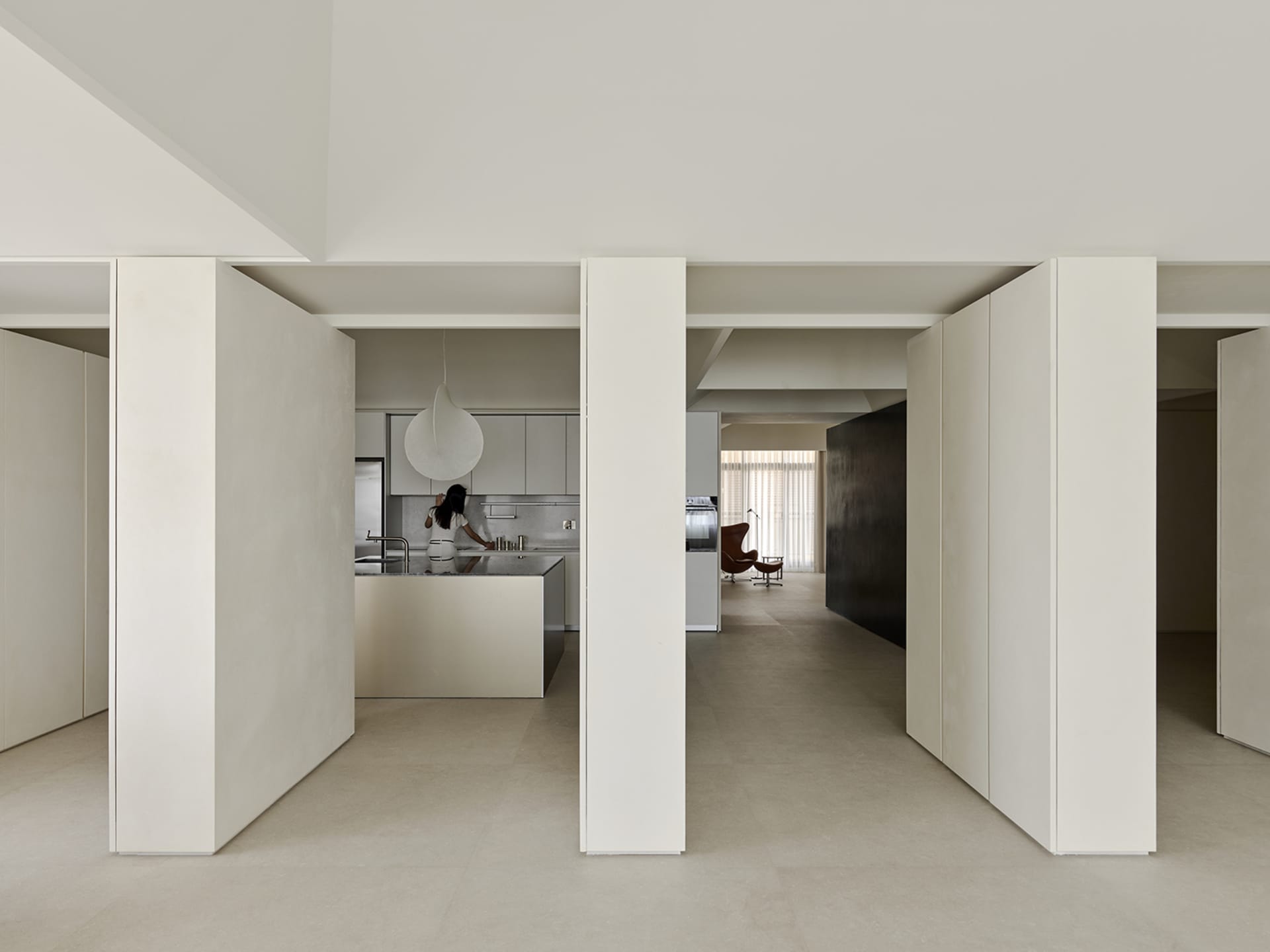 View from a hallway into a minimalist open-plan apartment featuring micro-cement walls, a central kitchen island, and a pendant light, with a woman cooking in the background.