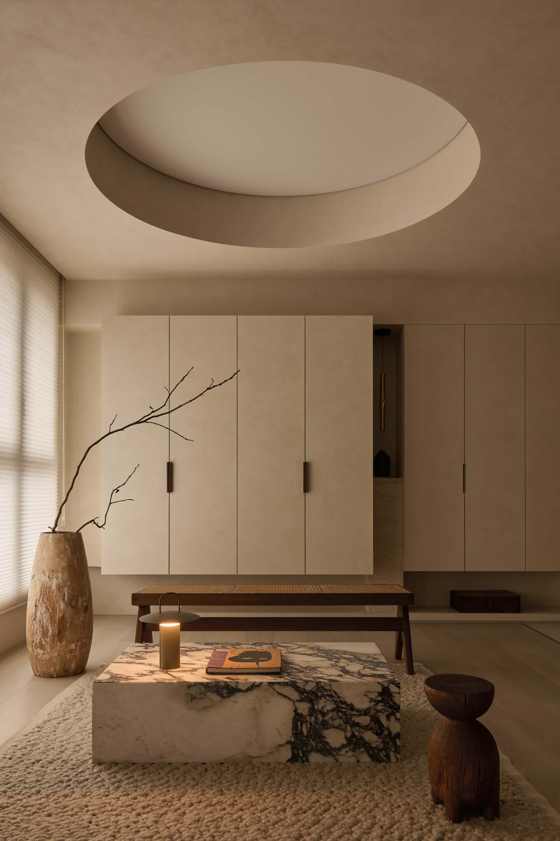 Minimalist living room featuring a large Calacatta marble coffee table, low wooden bench, sculptural dark wood stool, textured beige rug, built-in neutral cabinetry, and a dramatic circular recessed ceiling light.