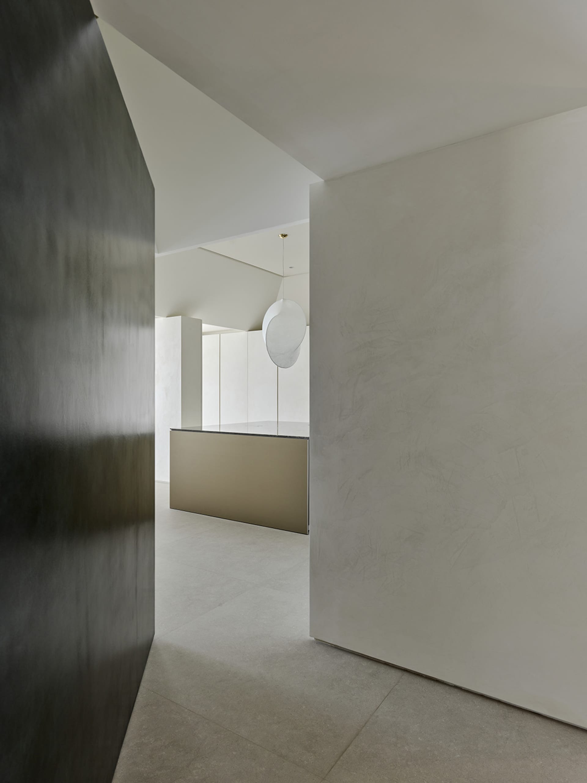 View from a dark hallway into a bright minimalist kitchen featuring micro-cement walls, concrete floor tiles, and a large white sculptural pendant lamp hanging over a metallic island.