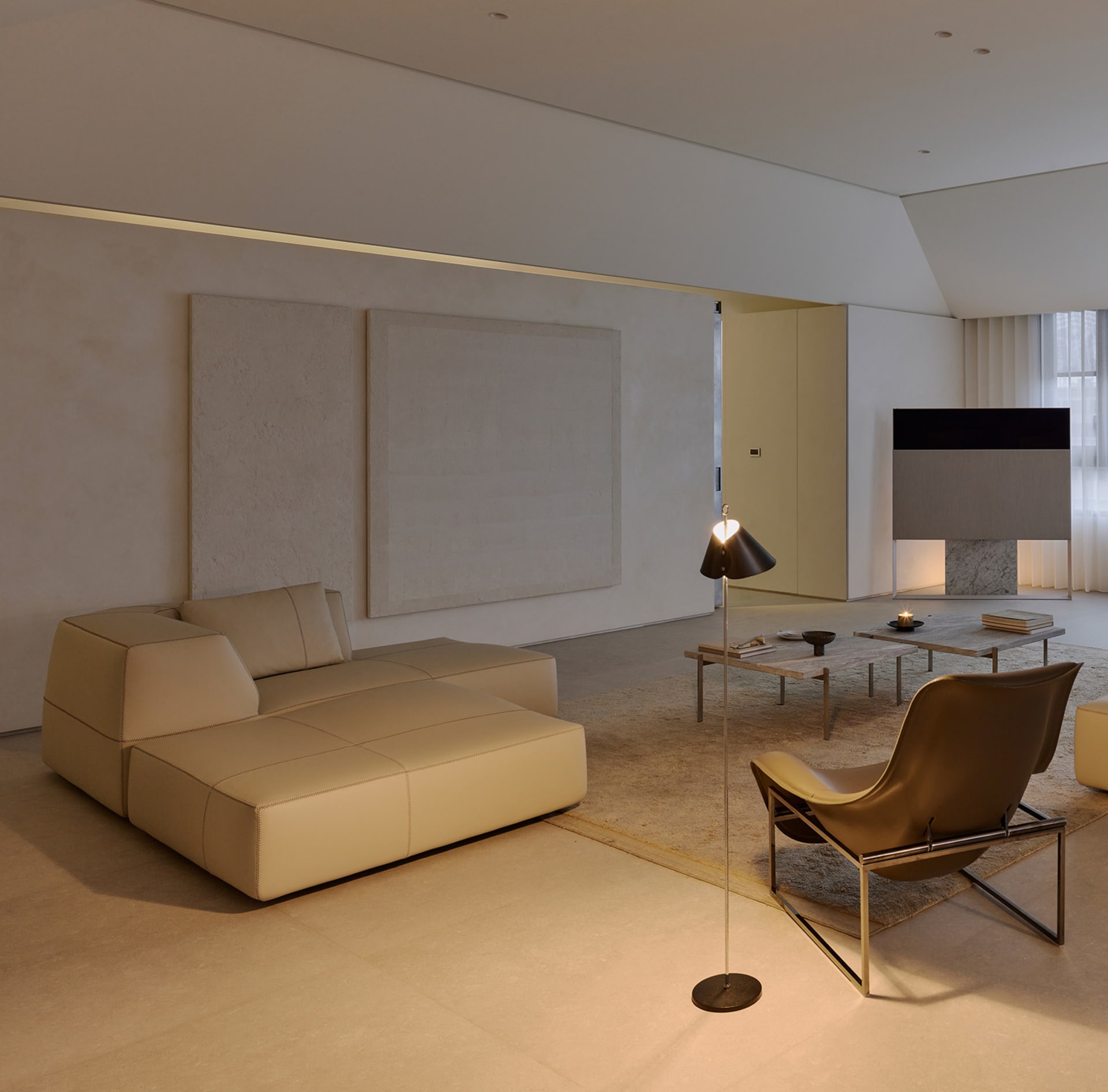 Minimalist living room featuring a large beige modular sofa, brown leather lounge chair, floor lamp, and expansive micro-cement walls with warm ambient lighting.