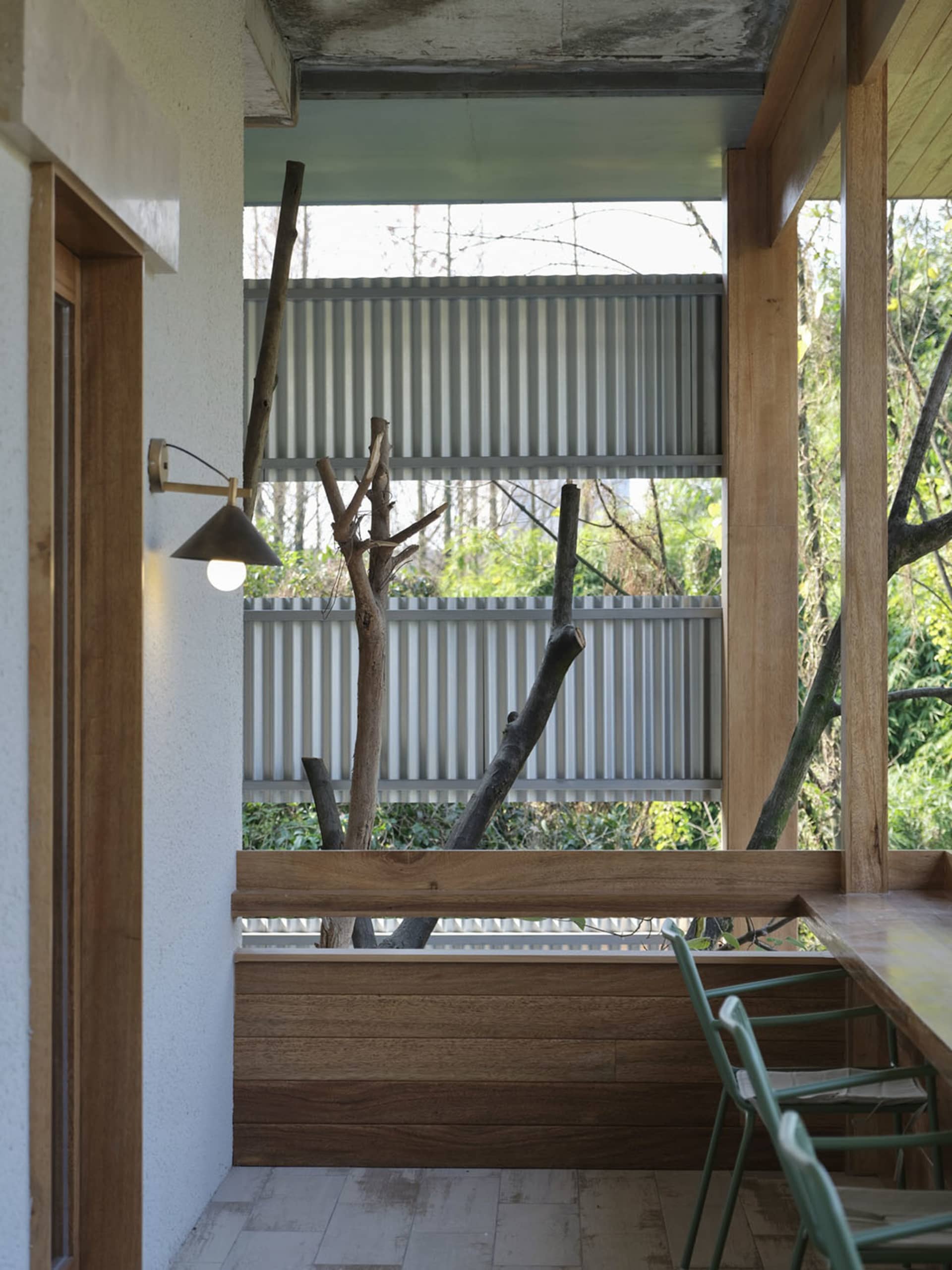 Covered patio interior featuring wooden walls, a large branch sculpture, corrugated metal panels, and green metal dining chairs overlooking trees.