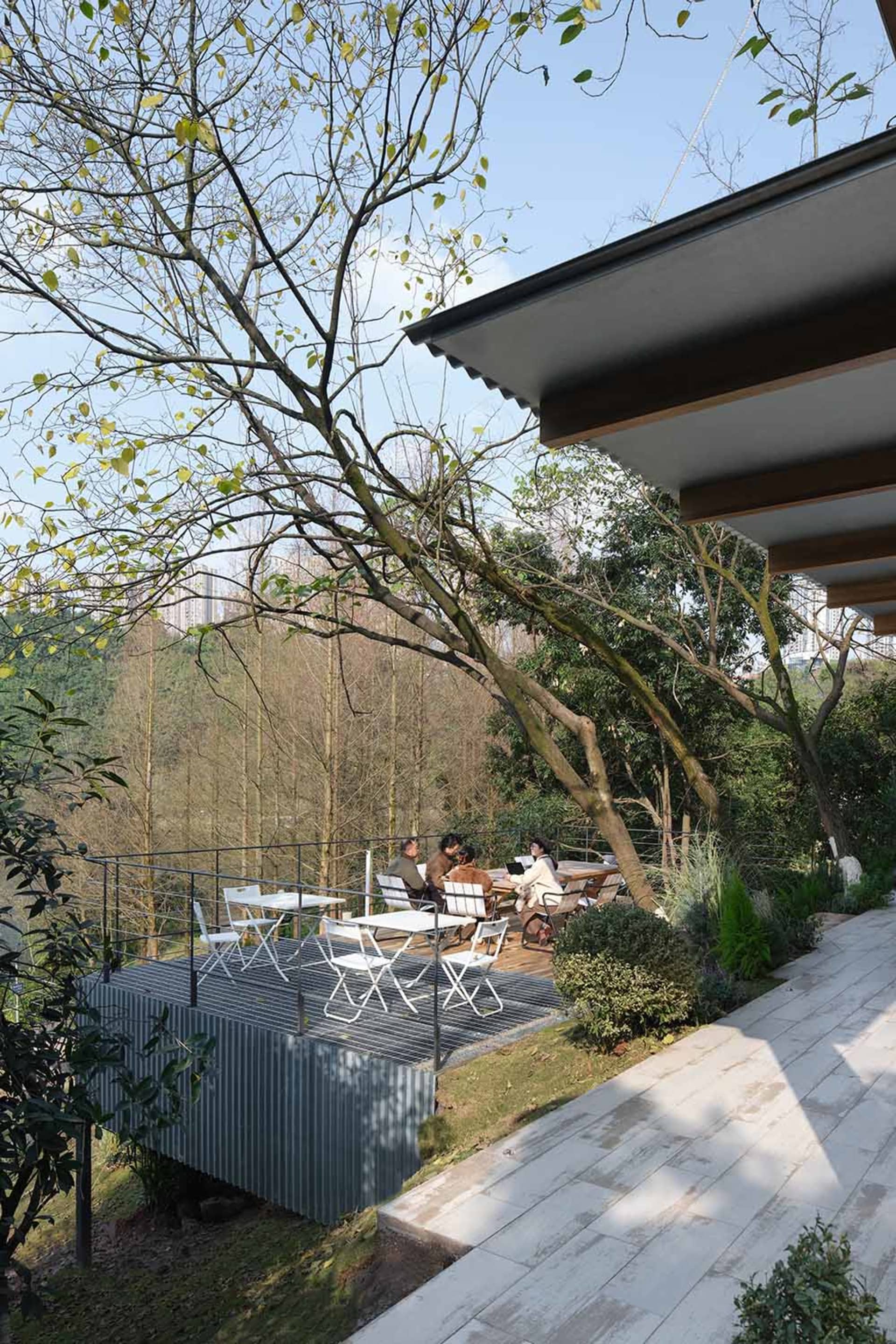 Modern outdoor deck with corten steel structure and white dining furniture overlooking a lush forested mountain valley, with people enjoying a meal under the trees.