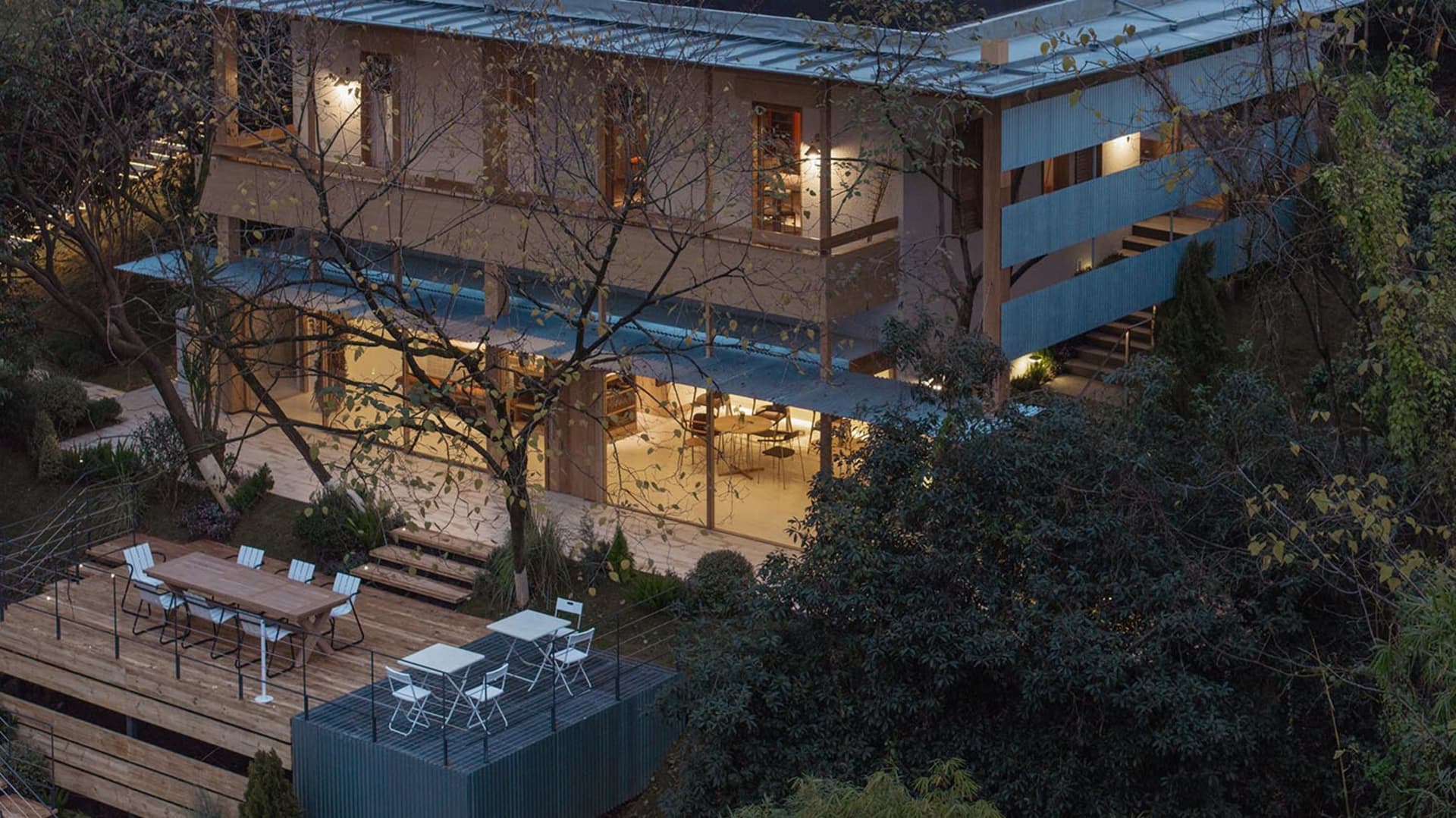 現代兩層別墅夜景,包含大面積木露台、下層耐候鋼貨櫃式結構搭配白色戶外家具,以及周圍茂密樹木。