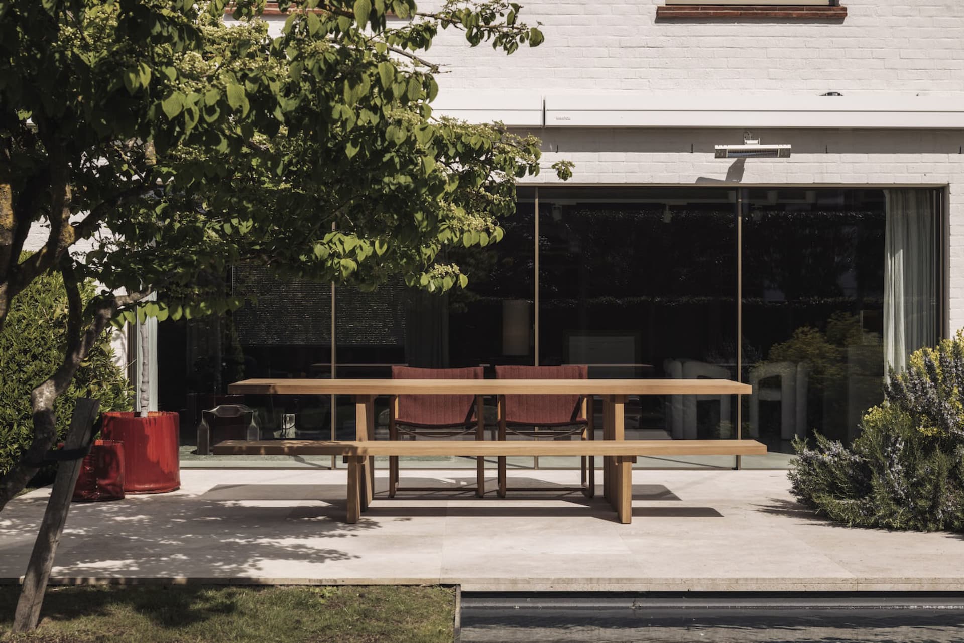 Modern outdoor dining area featuring a long solid teak wood table, matching bench, and two red upholstered chairs on a large-format stone patio in front of a white brick house with large glass doors.