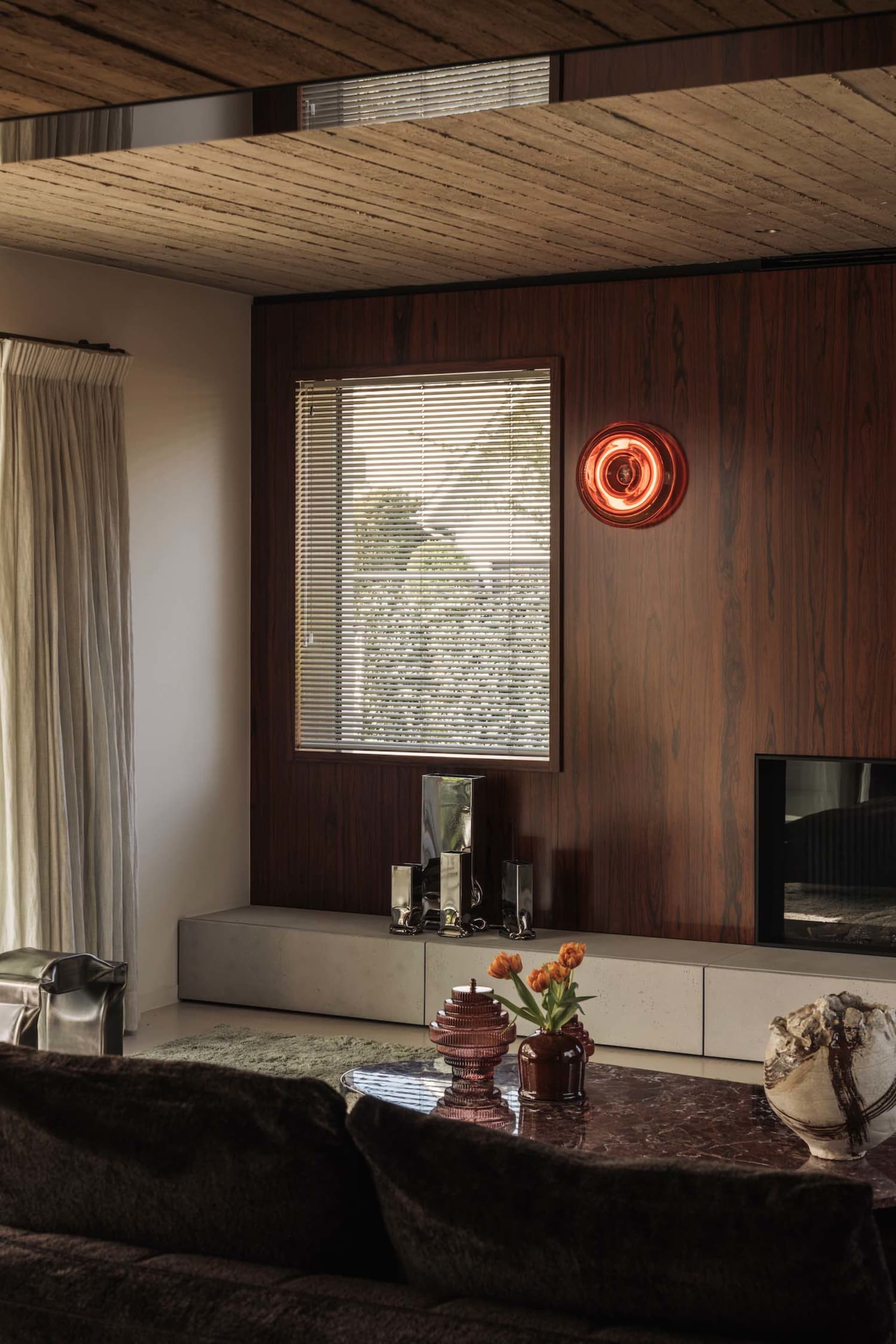 Modern living room featuring dark walnut wood paneling, a marble coffee table with glass and ceramic decor, a concrete media console, and warm ambient lighting from a red neon wall sconce.