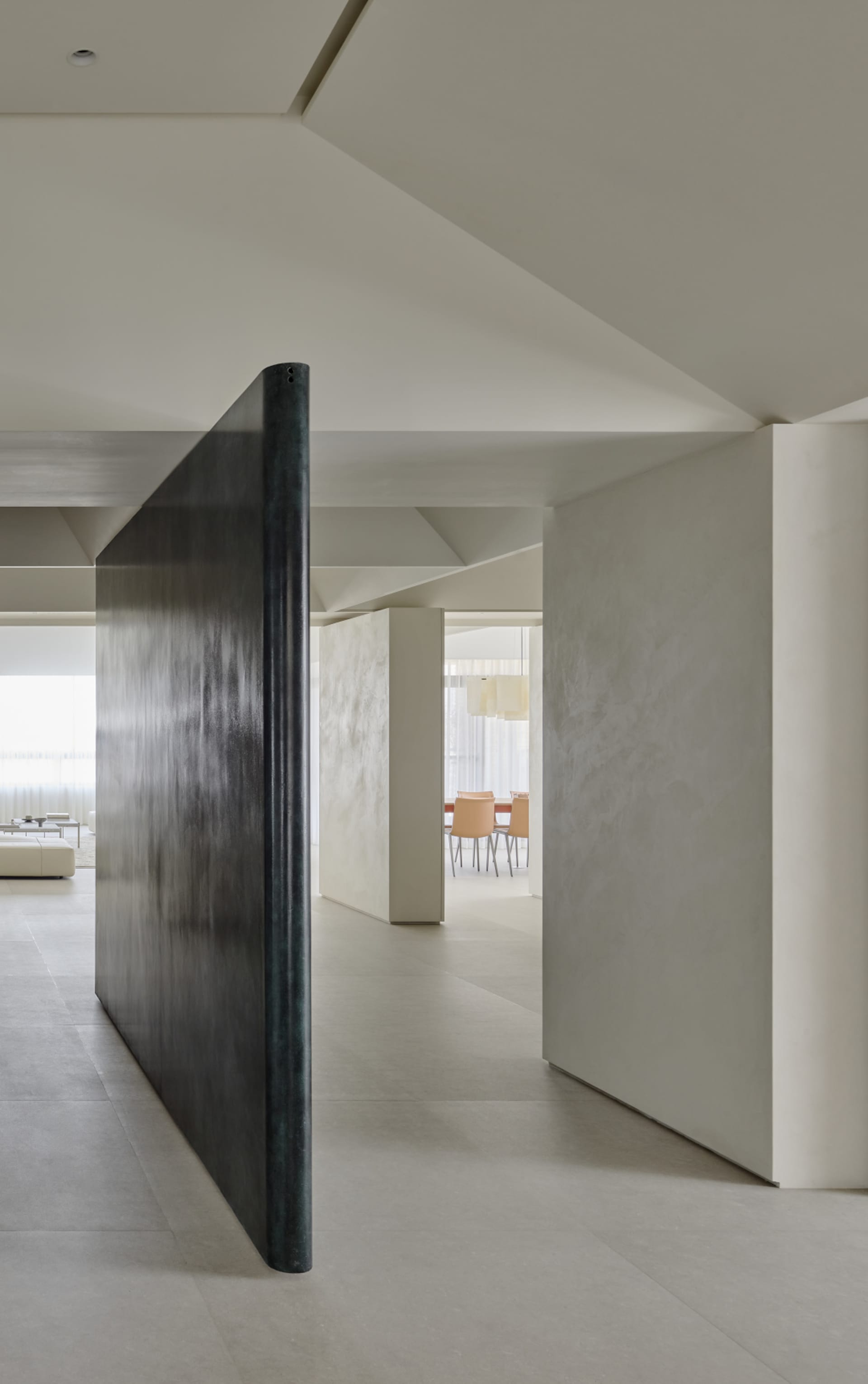 Modern minimalist interior featuring a tall patinated bronze pivot door leading to a bright open-plan living and dining area with neutral tones.