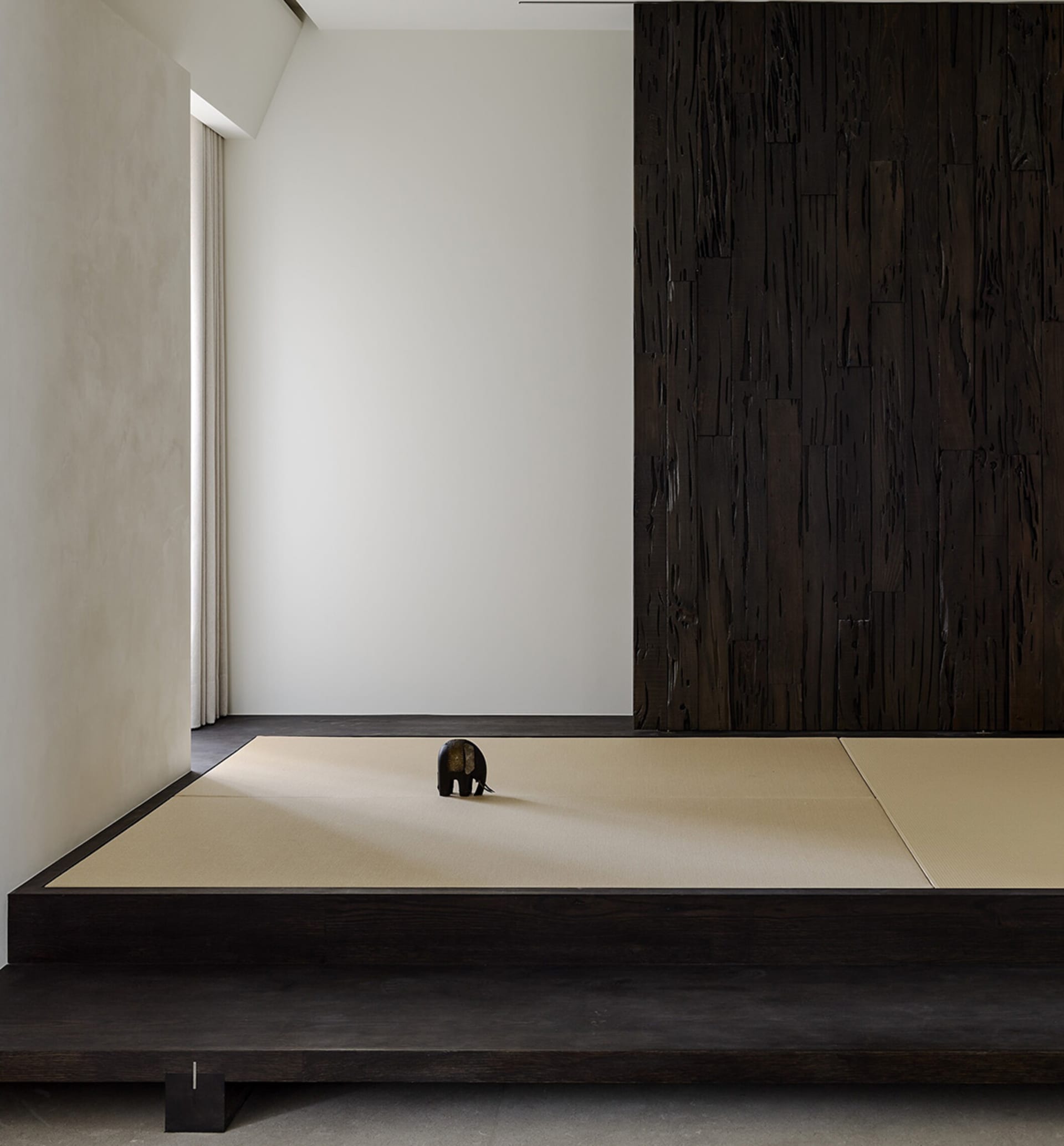 Minimalist Japanese-style room featuring a raised tatami mat platform, dark charred wood accent wall, light beige tatami mats, and a small dark elephant sculpture placed in the center.