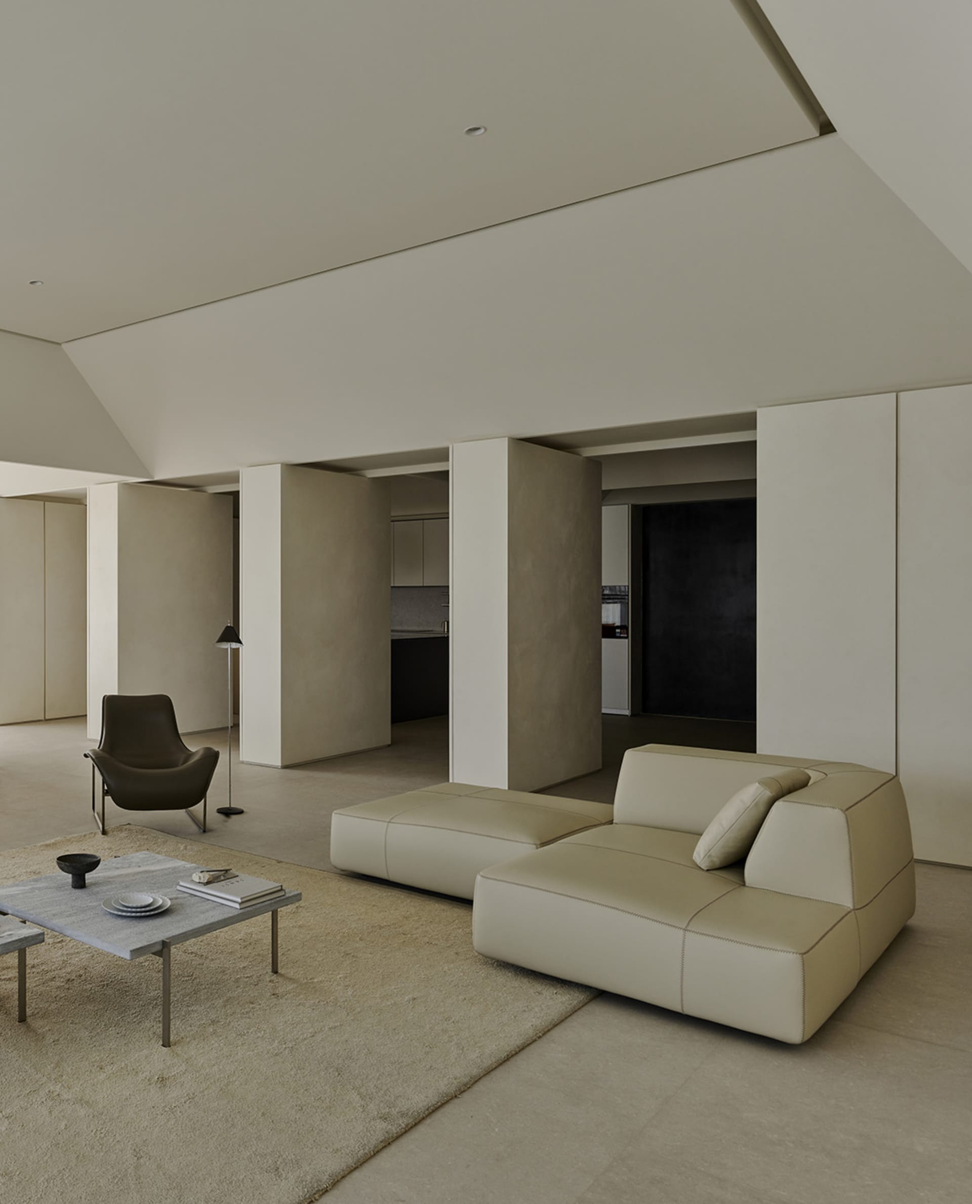 Minimalist living room featuring a large beige leather modular sofa, low concrete coffee table on a textured rug, dark leather lounge chair, and high concrete ceilings with soft natural lighting.